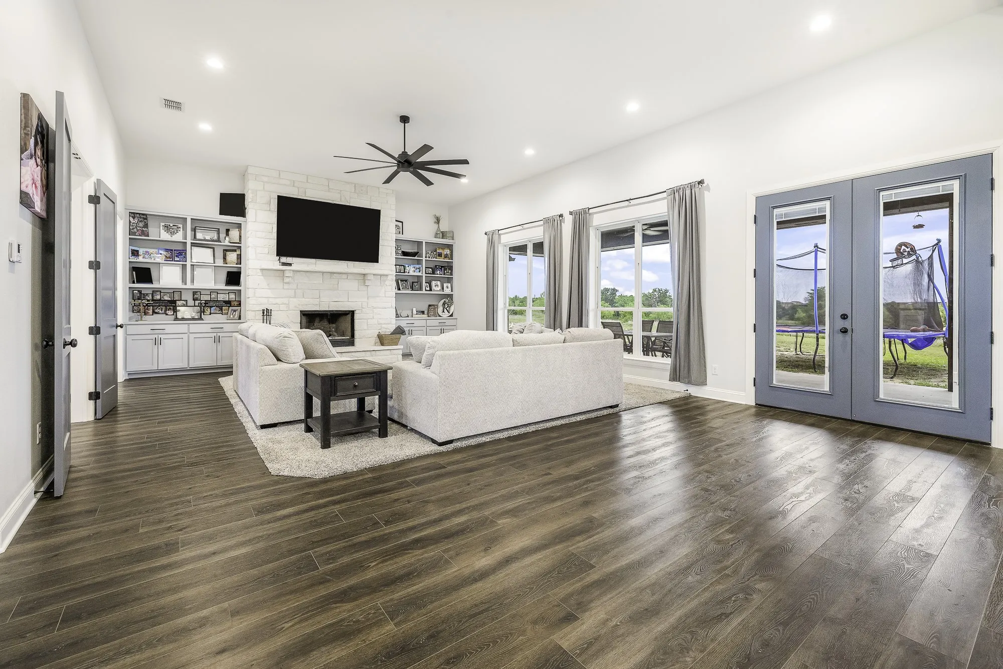Spacious living area featuring wood-finish flooring, a stone-clad fireplace, integrated shelving with lower cabinetry, and multiple large windows with exterior access