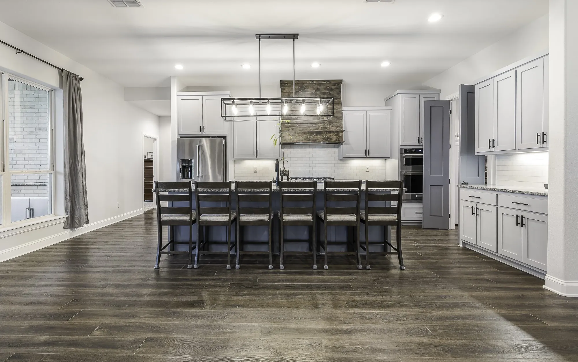 Open-concept kitchen featuring a large central island with seating, dark wood-finish flooring, white shaker cabinetry with dark hardware, stainless steel appliances, and a wood-wrapped range hood