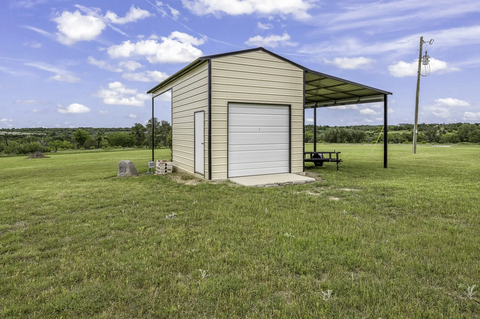 Metal utility building featuring a roll-up garage door, pedestrian entry door, covered carport extension, and concrete pad