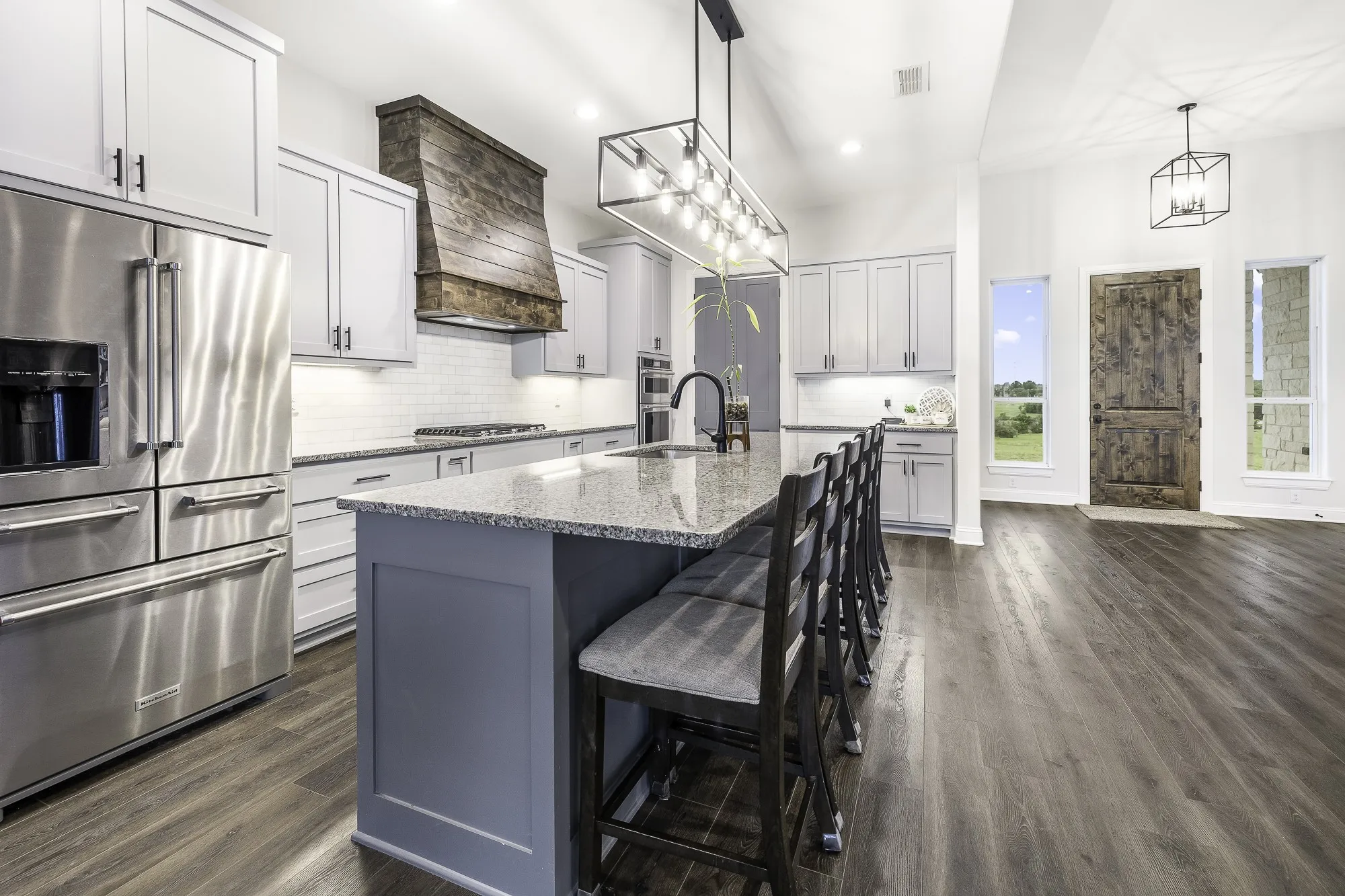 Open-concept kitchen featuring an oversized island with seating, stainless steel appliances, a wood-finish range hood, and white subway tile backsplash