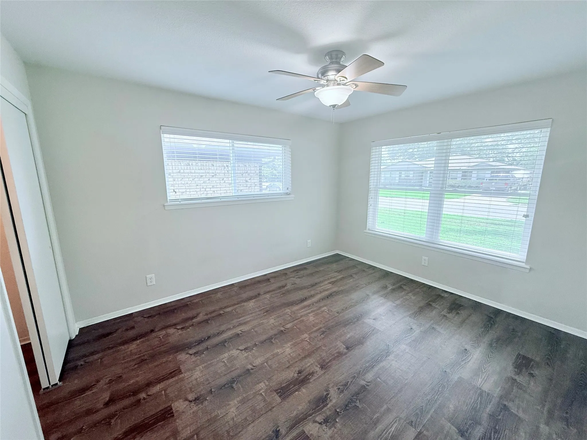 Spacious room featuring wood-finish flooring, two windows with blinds, a ceiling fan with integrated lighting, and light-toned walls