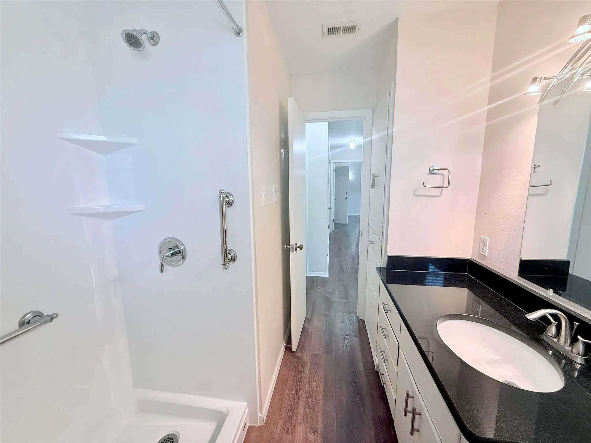 Bathroom featuring a white shower with grab bars and built-in shelving, a dark granite-style countertop with an undermount sink, a vanity with white cabinetry and brushed nickel hardware, and wood-finish flooring
