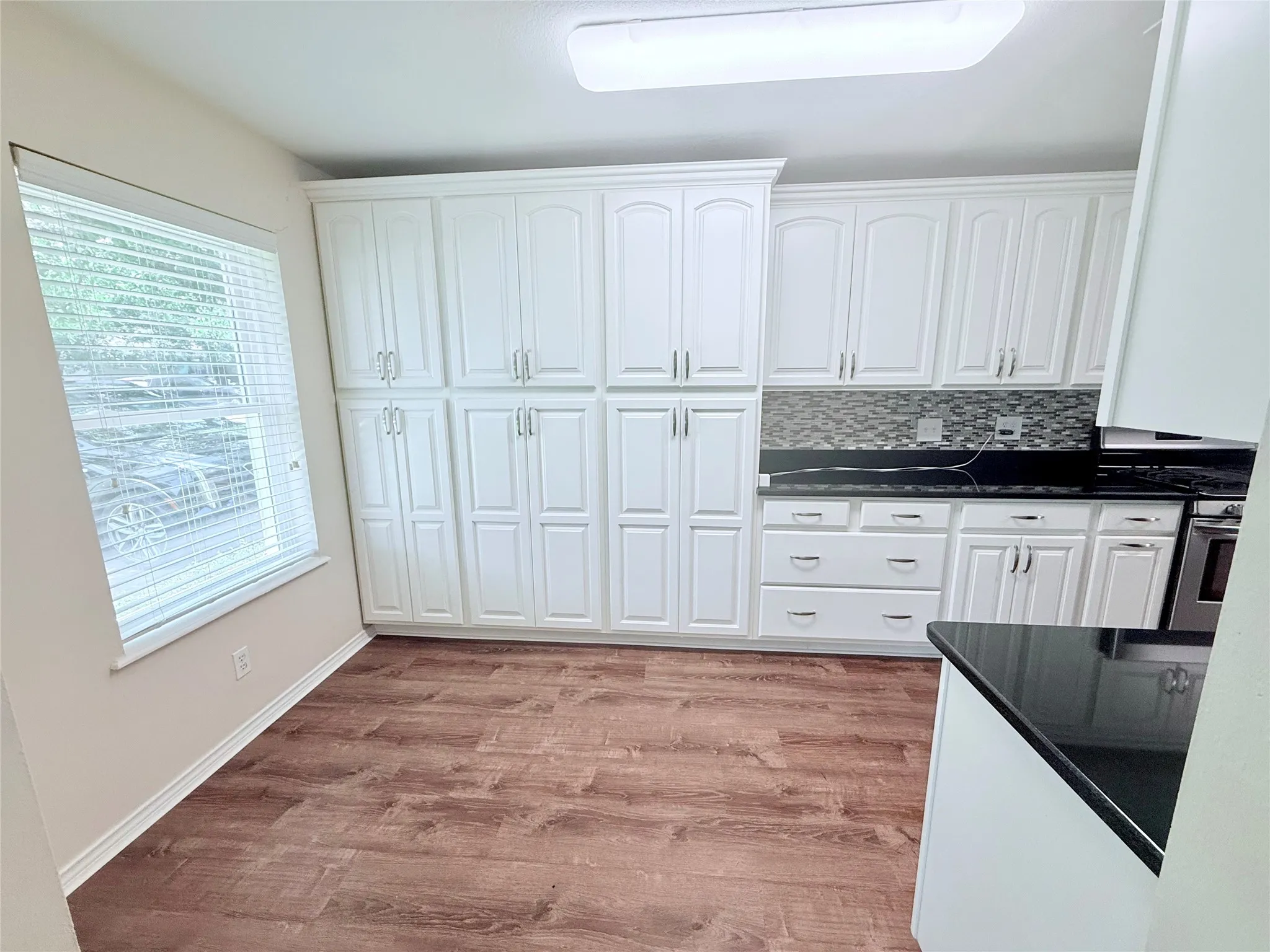 Kitchen featuring extensive white cabinetry, dark countertops, wood-finish flooring, a mosaic tile backsplash, and a window with blinds