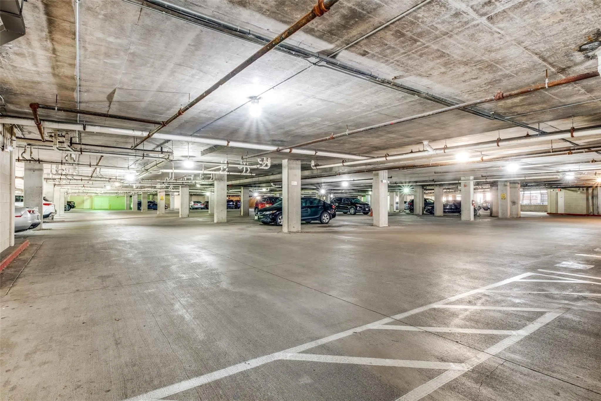 Spacious concrete parking garage with multiple columns, overhead piping, and ceiling-mounted lighting fixtures
