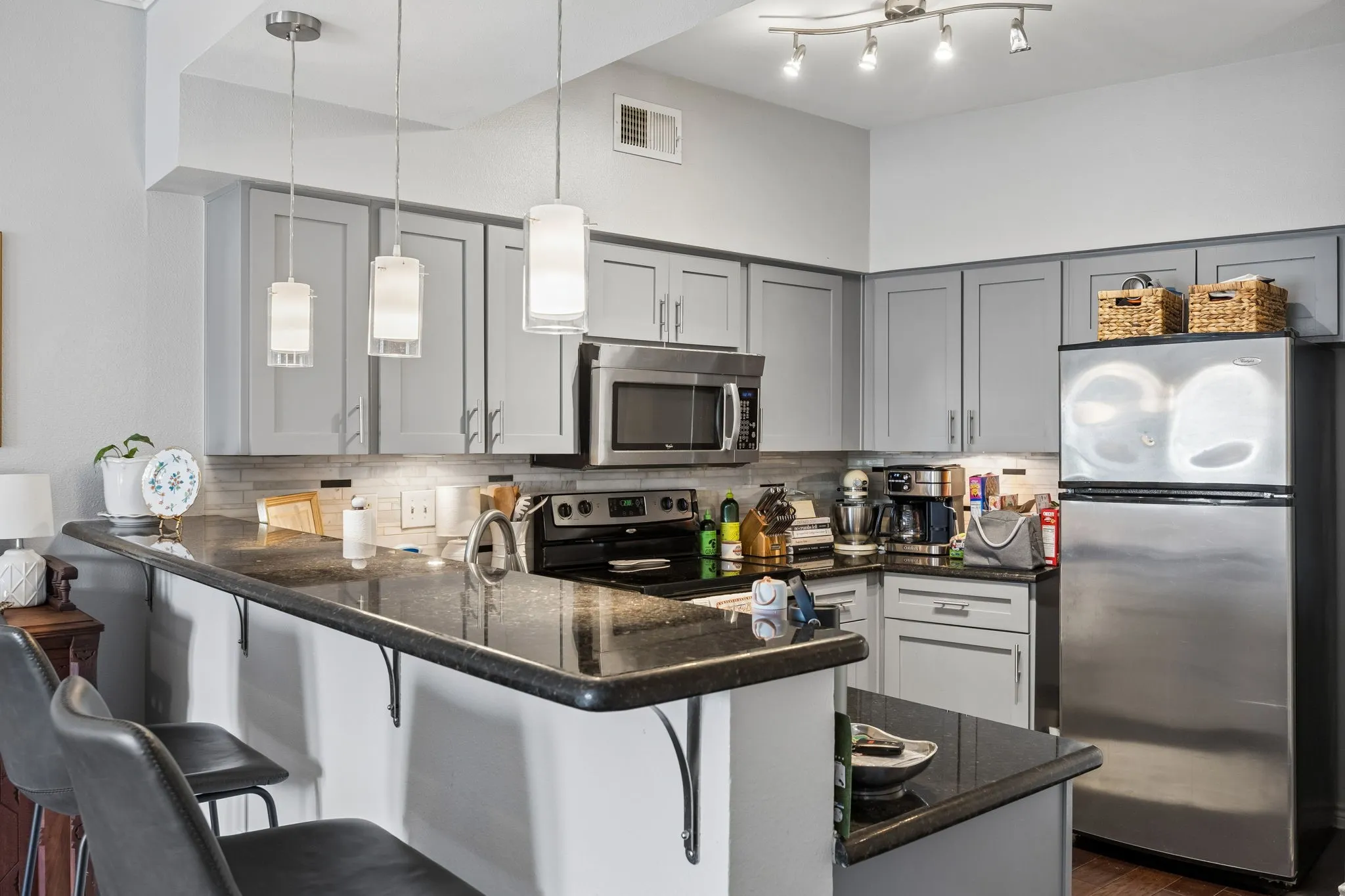 Kitchen featuring gray cabinetry, stainless steel appliances, dark stone countertops, a tile backsplash, and pendant lighting over a breakfast bar