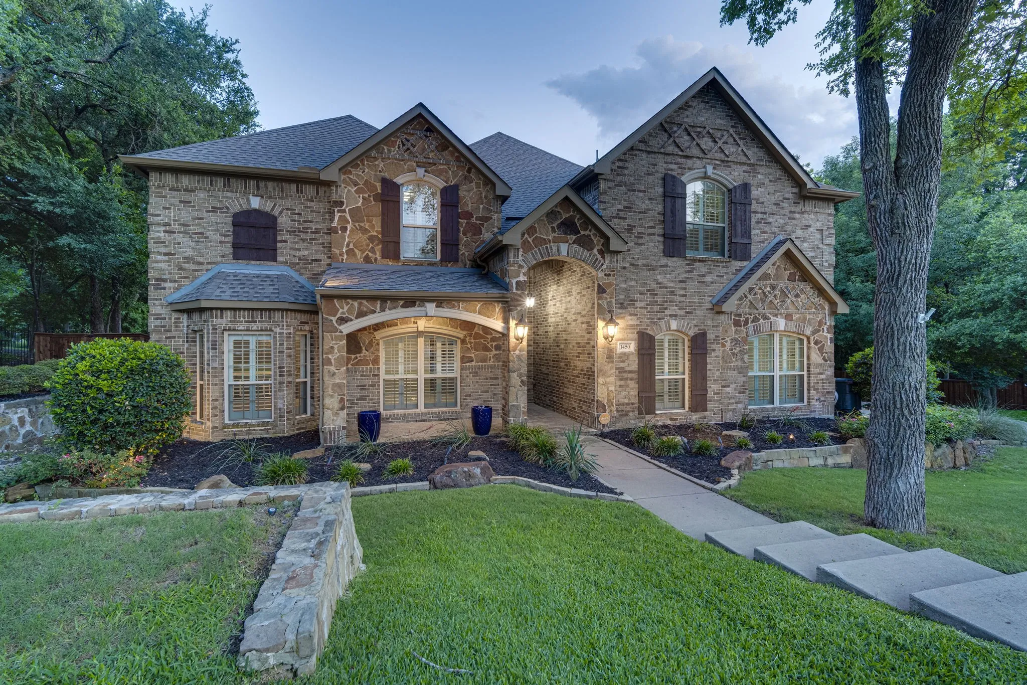 Front elevation framed by mature trees and manicured landscaping at twilight viewing .