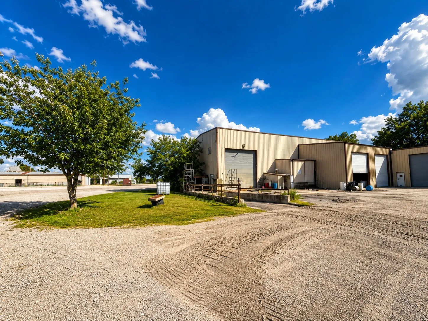 Industrial building with multiple bay doors, gravel lot, and partial grassy area with mature trees