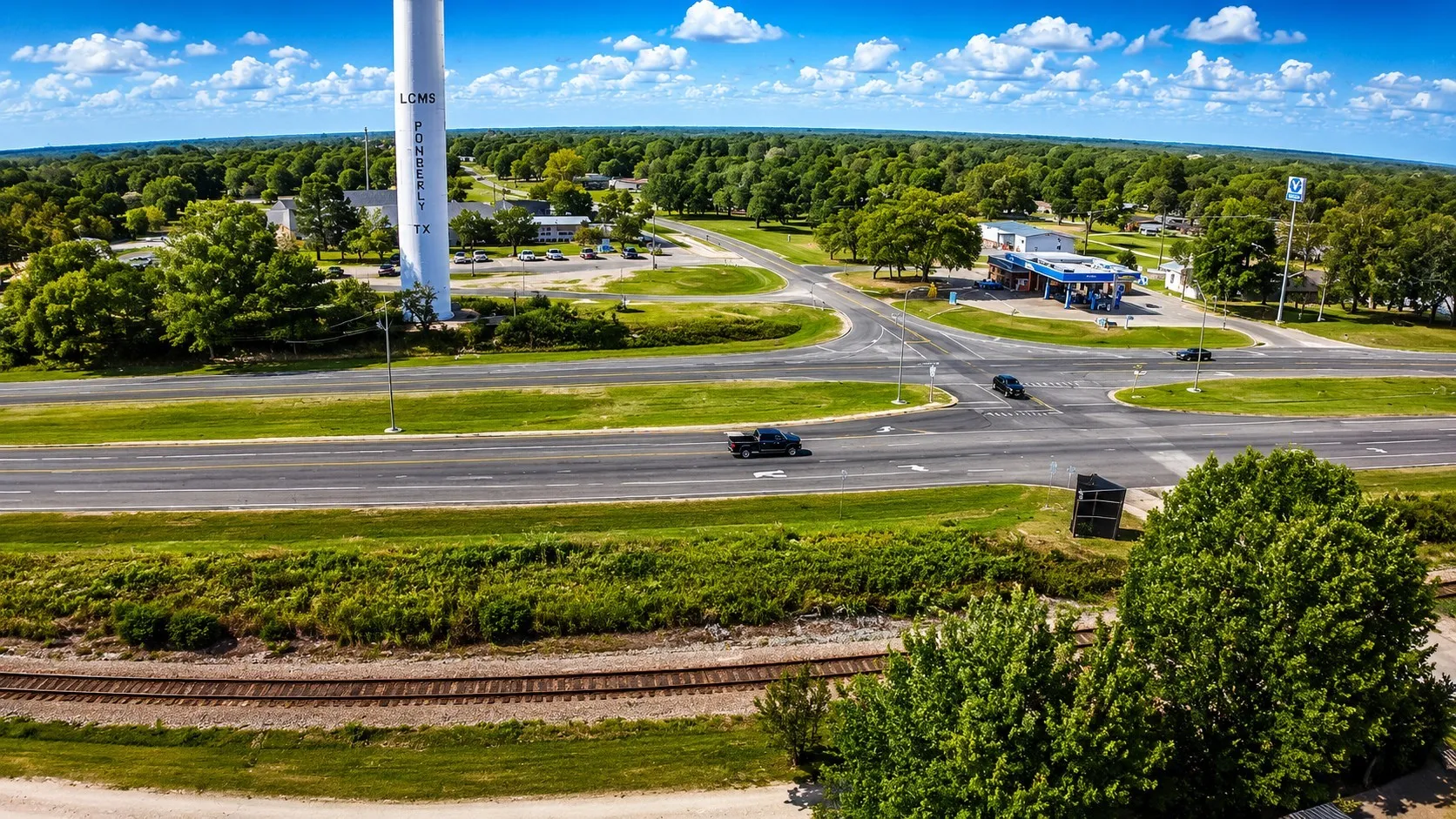 Aerial perspective of a commercial intersection with multiple businesses, including a gas station, surrounded by mature trees