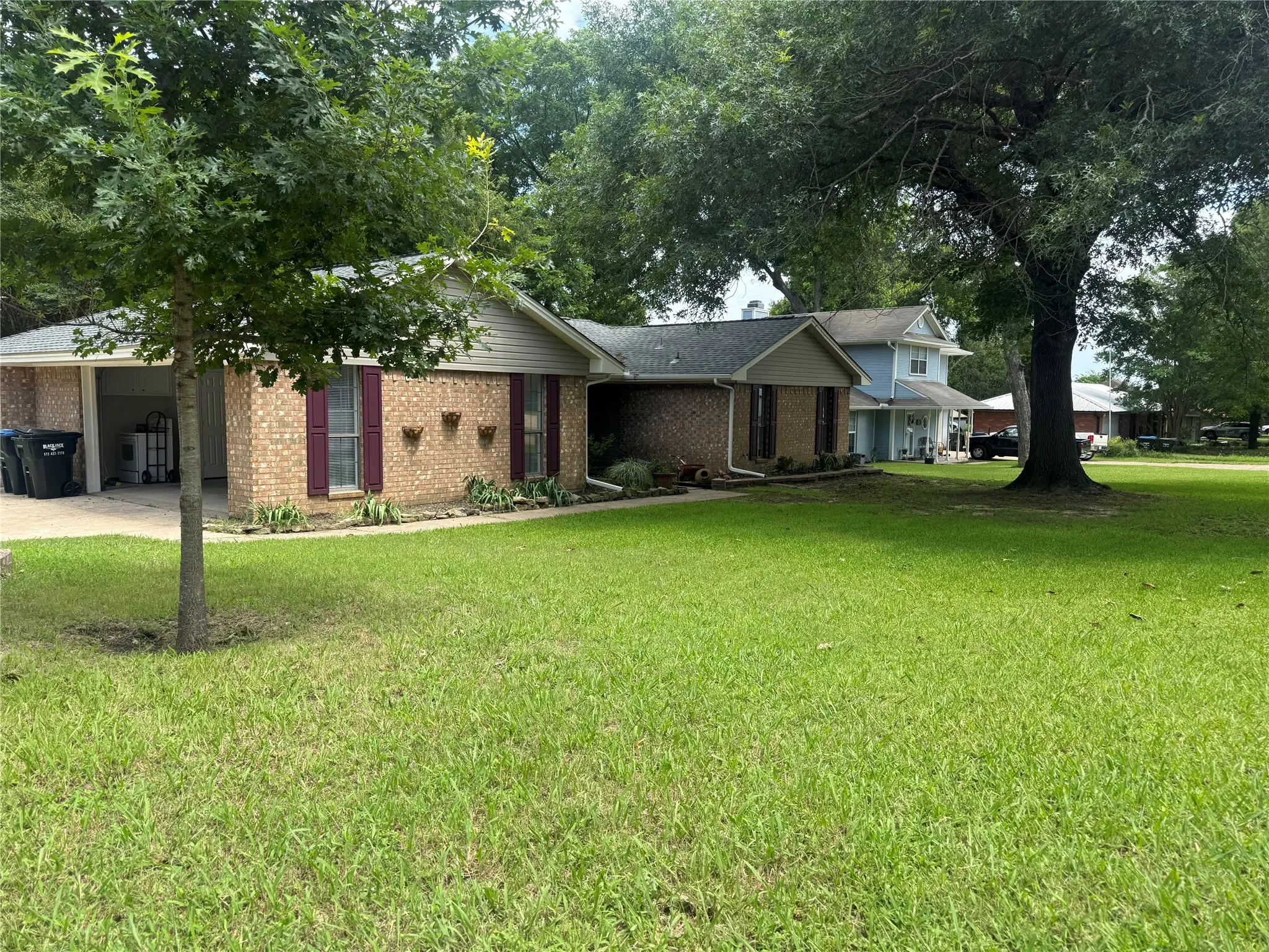 Brick exterior featuring dark red shutters, a prominent tree, a lush green lawn, and a light-colored roof