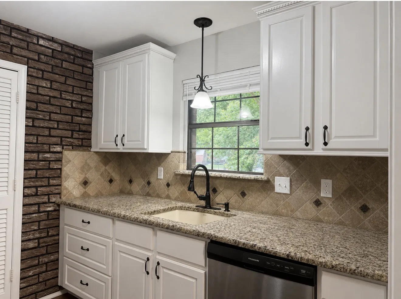 Kitchen featuring granite countertops, white cabinetry with dark hardware, a tile backsplash with accent squares, a stainless steel dishwasher, and a brick-look accent wall