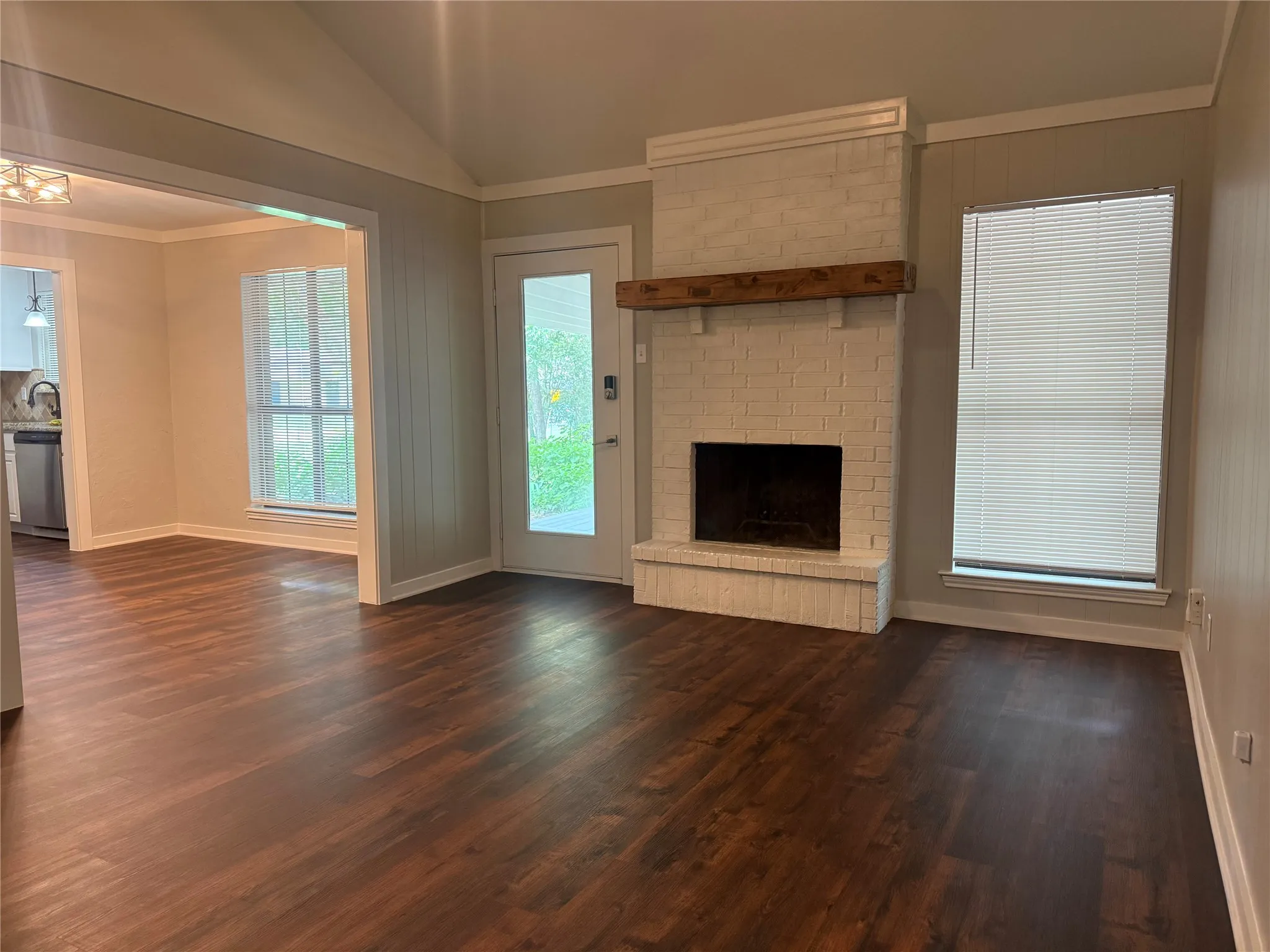 Spacious living area featuring wood-finish flooring, a white-painted brick fireplace with a wood mantle, and vaulted ceilings