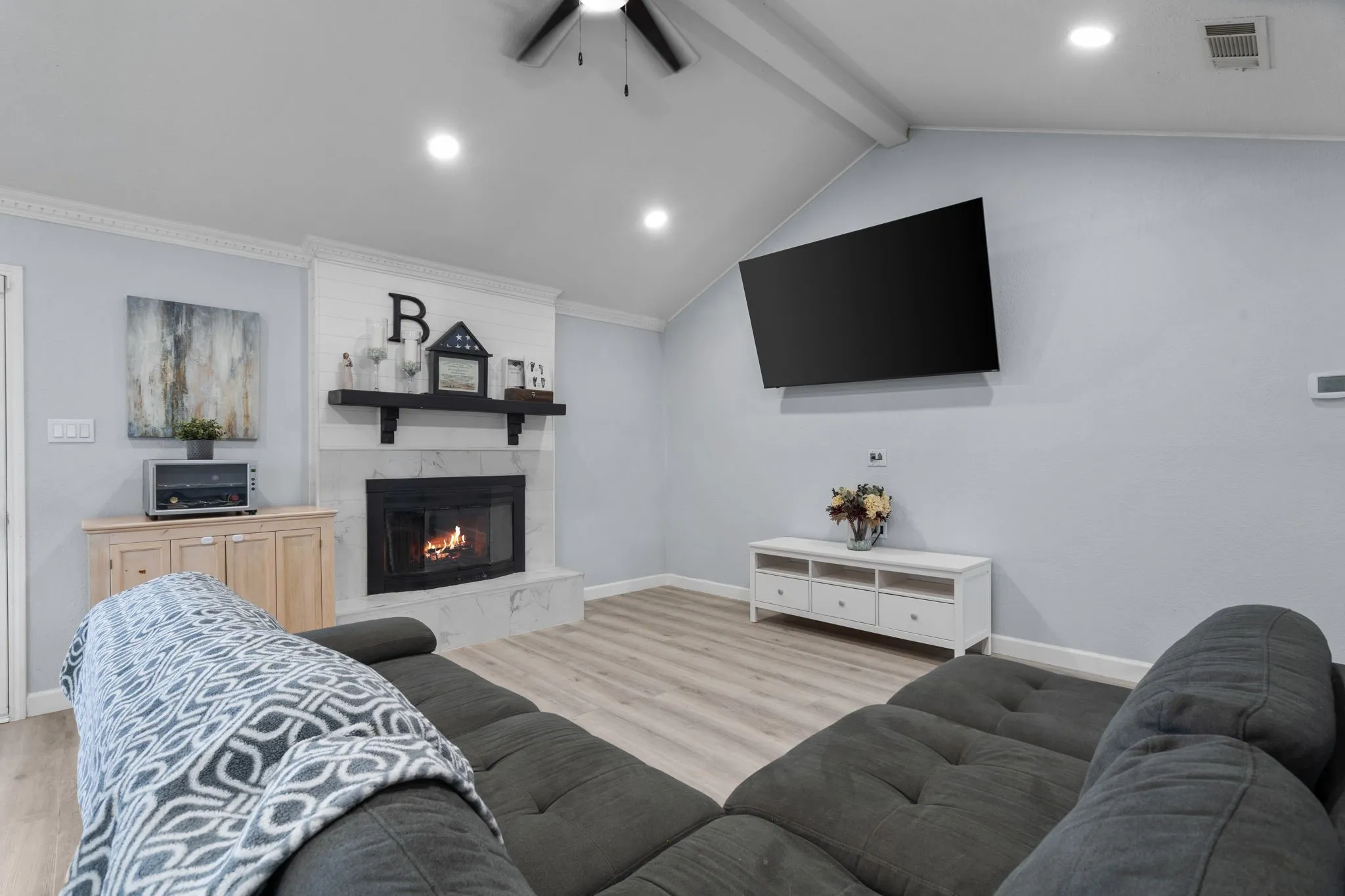 Vaulted ceiling living area featuring a central fireplace with a dark mantel, light-toned wood-finish flooring, and recessed lighting
