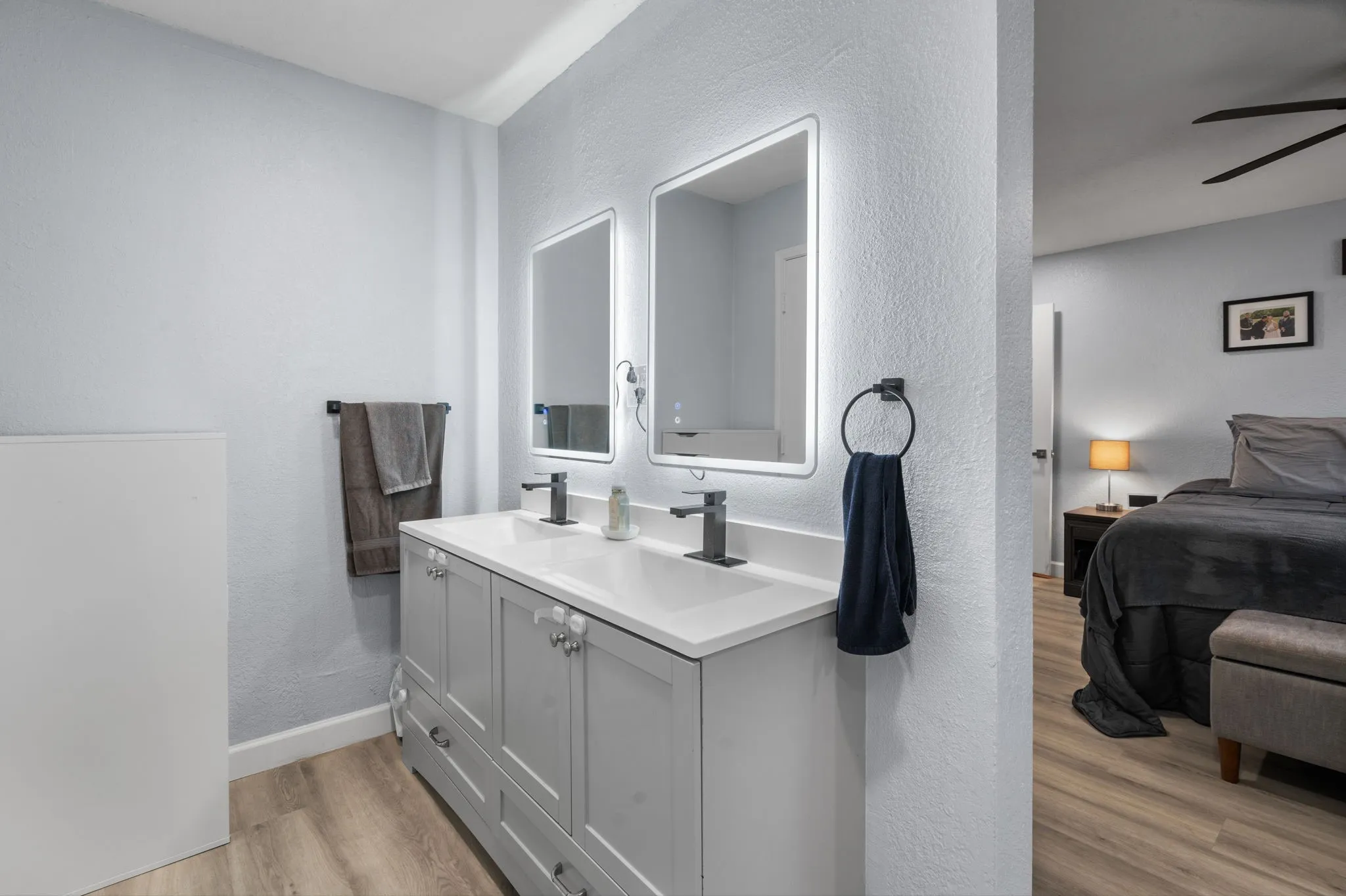 Dual vanity with integrated sinks and white countertops, illuminated mirrors, matte black fixtures, and wood-finish flooring