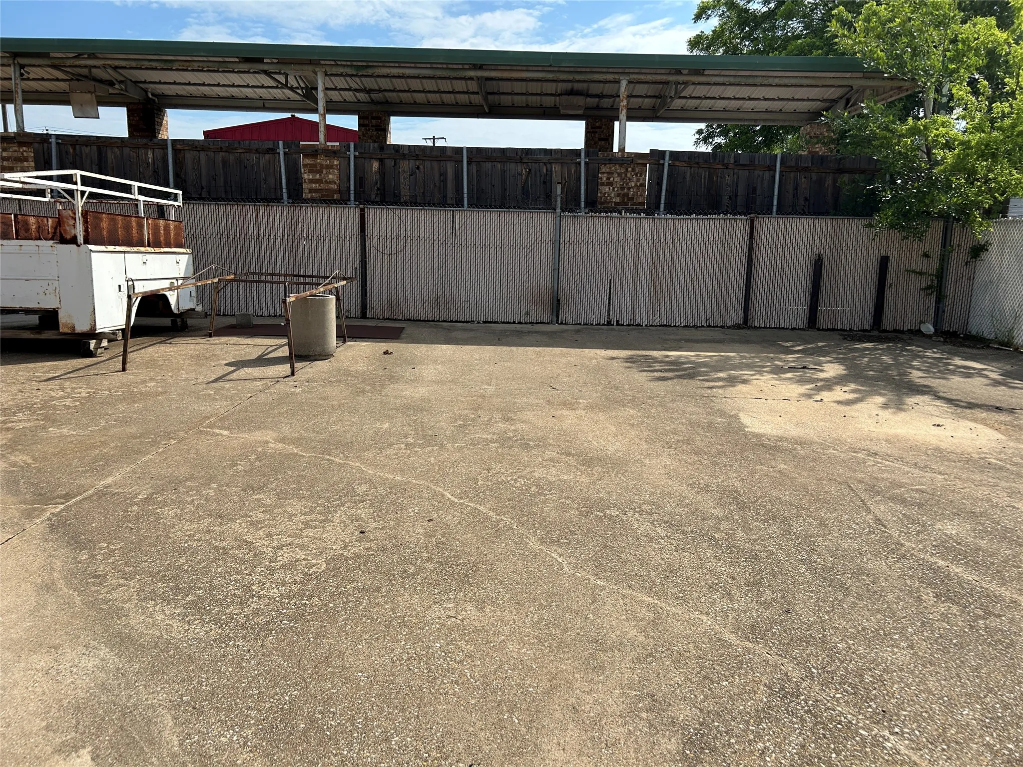 Paved concrete lot featuring a partial metal awning structure, chain-link fencing, and a wood privacy fence