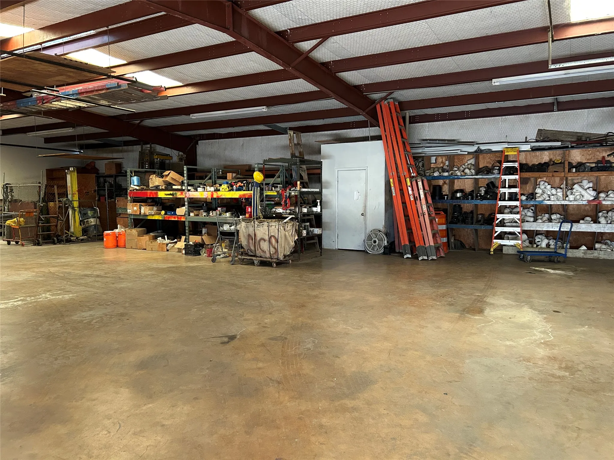 Spacious industrial interior featuring a high ceiling with exposed structural beams, ample open floor space, polished concrete flooring, fluorescent lighting, and a private enclosed room with a solid door