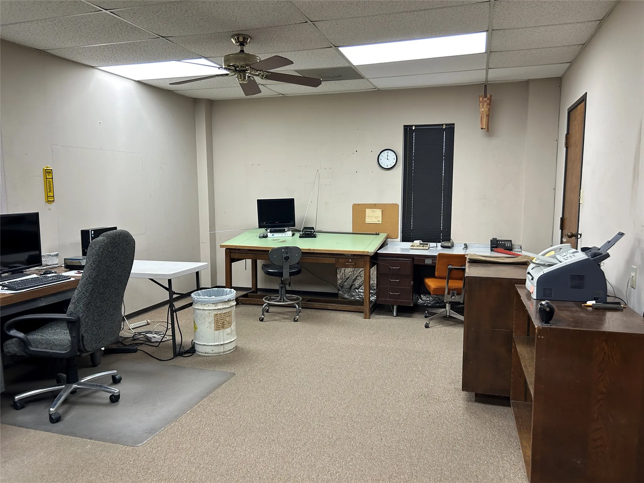 Commercial office space featuring textured carpet flooring, dropped acoustic ceiling tiles, recessed fluorescent lighting, a ceiling fan, and a solid core entry door