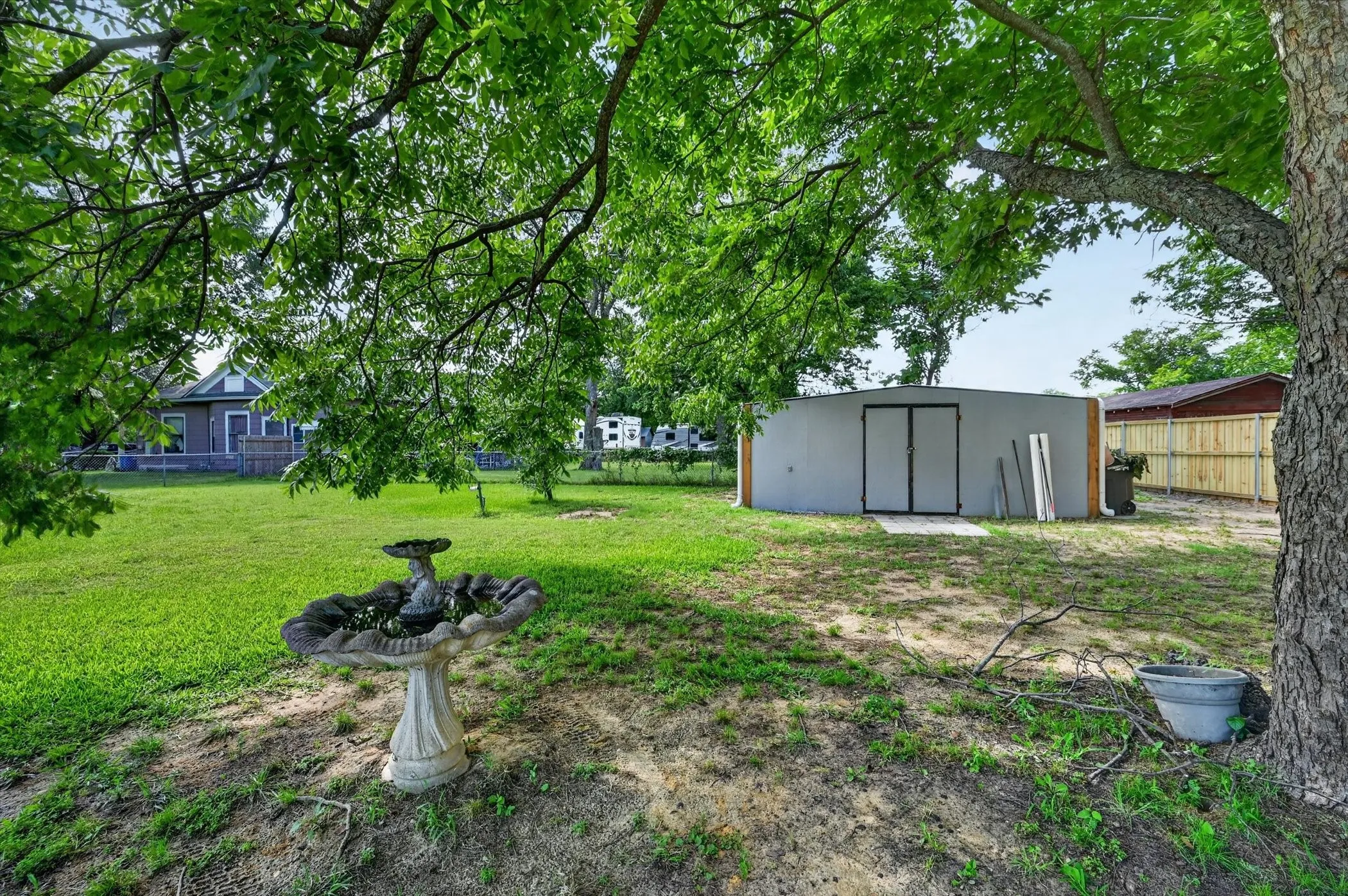 Expansive green space featuring mature trees, a decorative bird bath, a detached utility building with double doors, and a wood fence