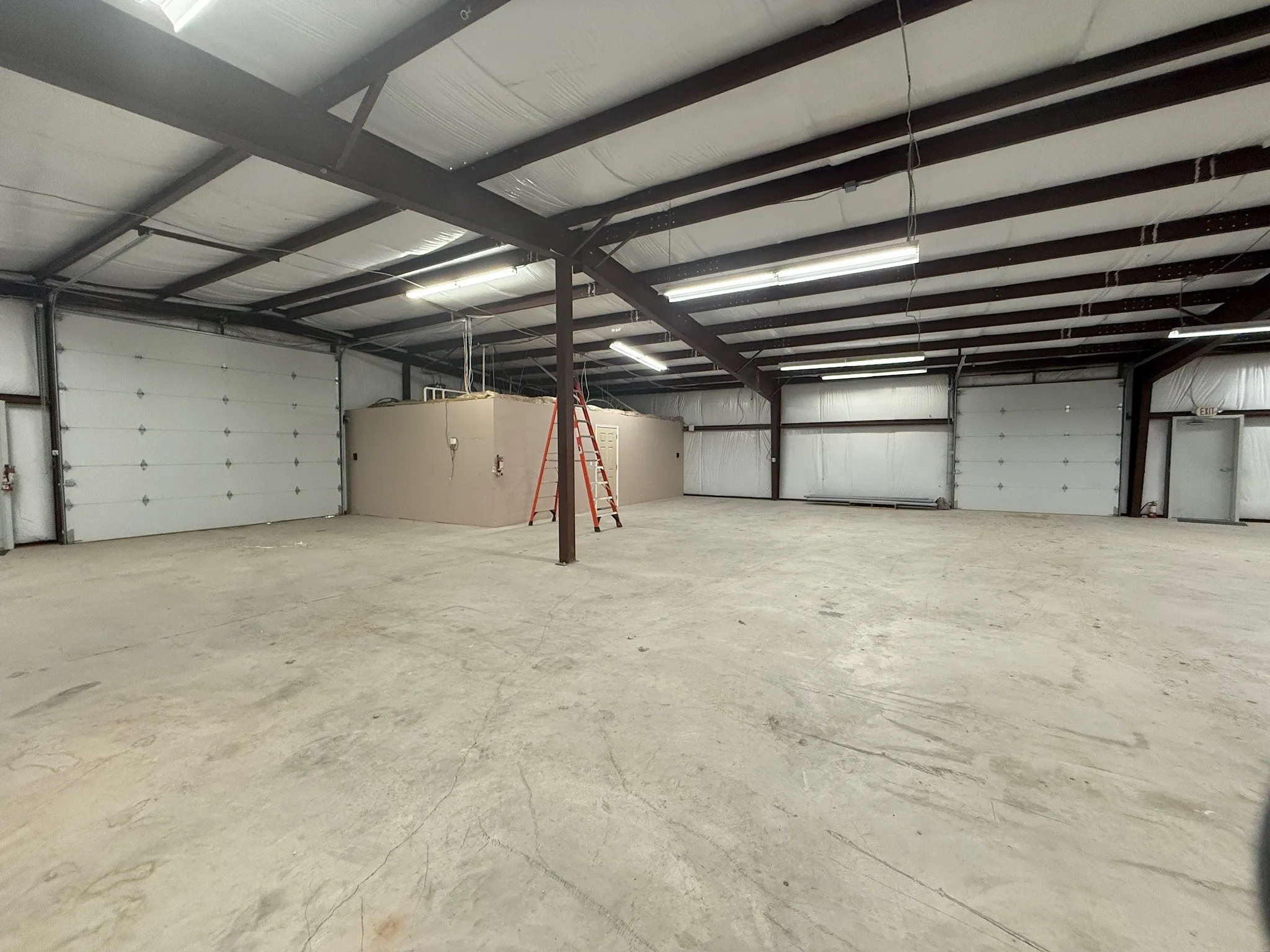 Spacious industrial interior featuring exposed steel I-beams, multiple overhead fluorescent lighting fixtures, and a concrete floor