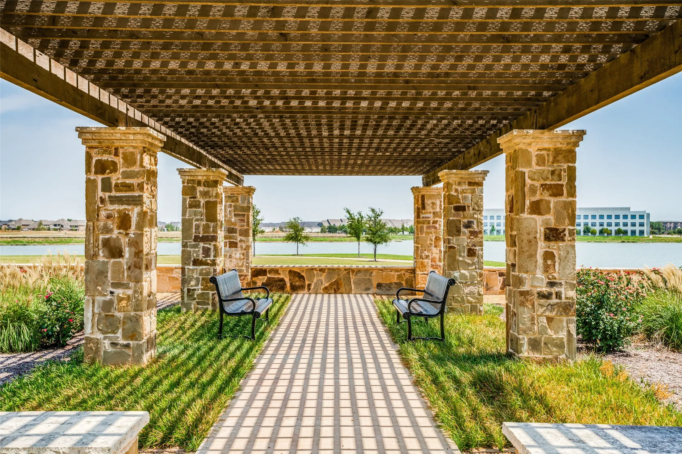 Stone-clad pergola structure with a slatted roof, featuring a paved pathway and flanking green spaces