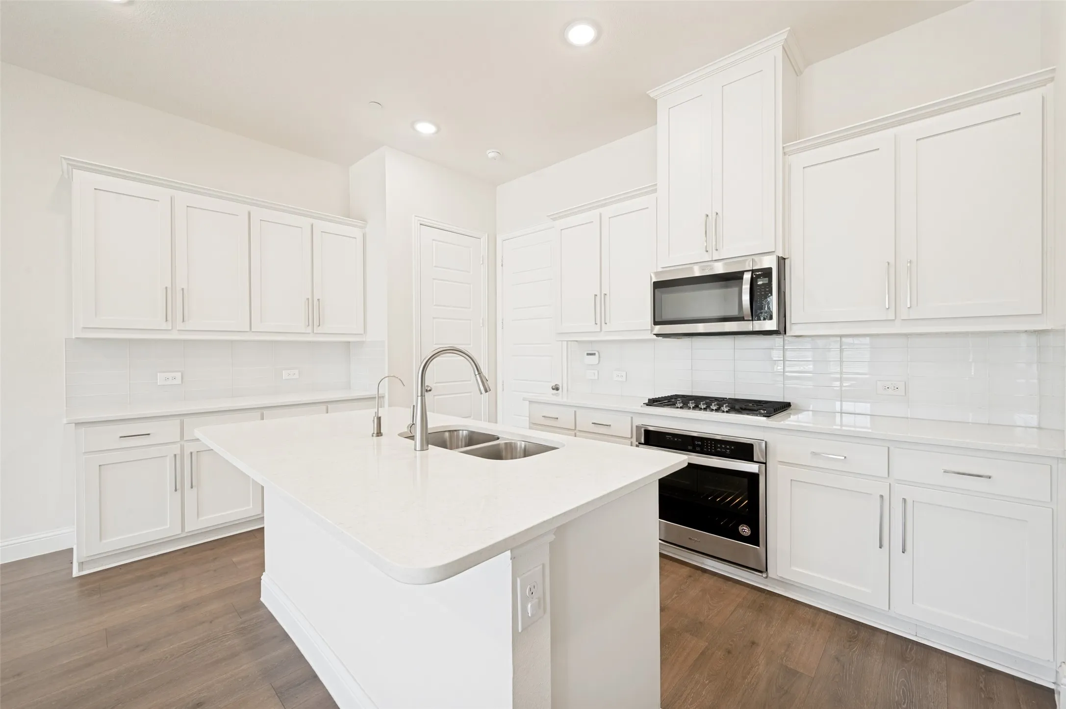 Modern kitchen featuring a central island with an undermount double-basin sink, white cabinetry, stainless steel appliances, white subway tile backsplash, and wood-finish flooring