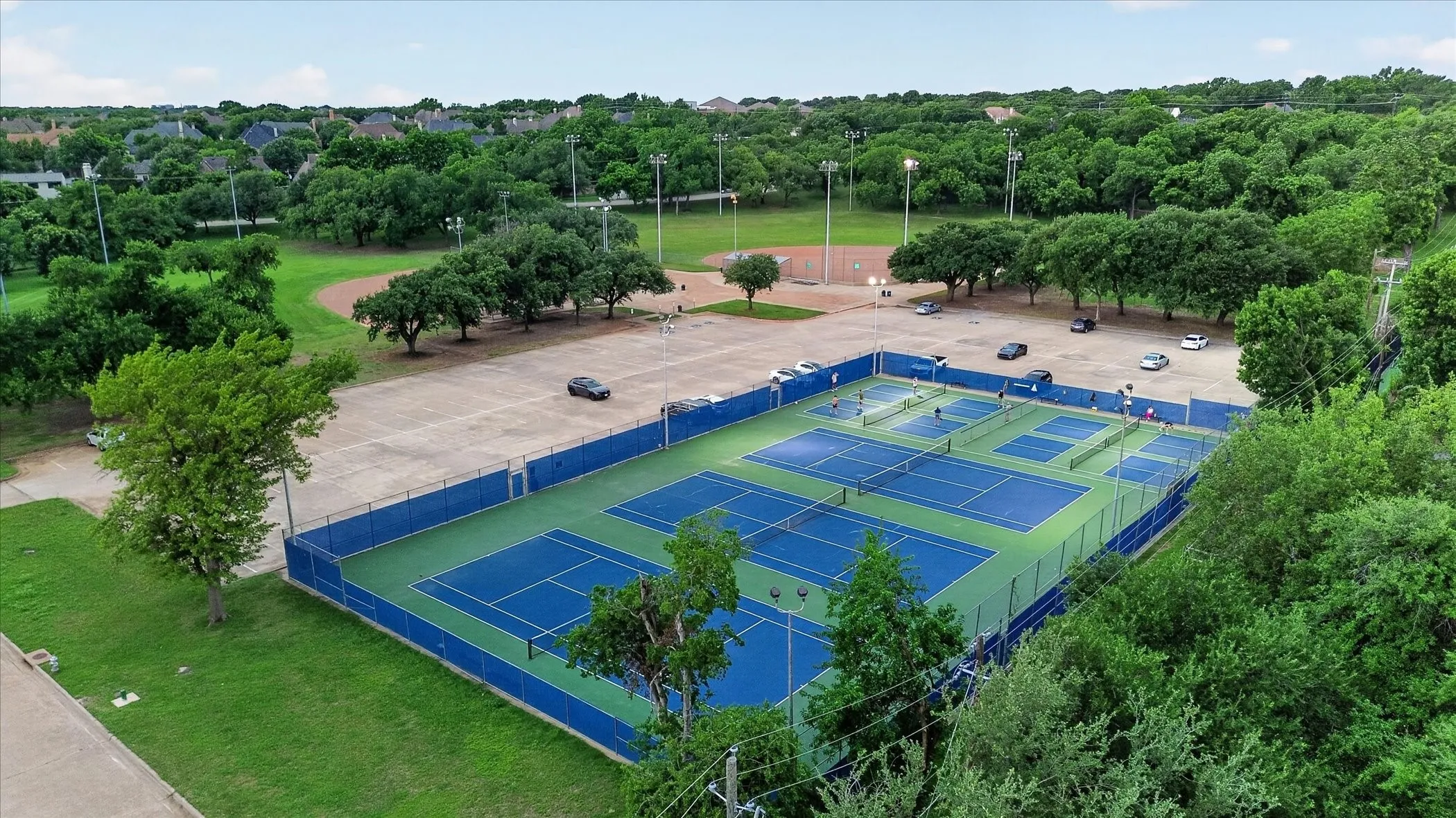 Multi-court recreational facility featuring vibrant blue and green playing surfaces, surrounded by mature trees and paved parking areas