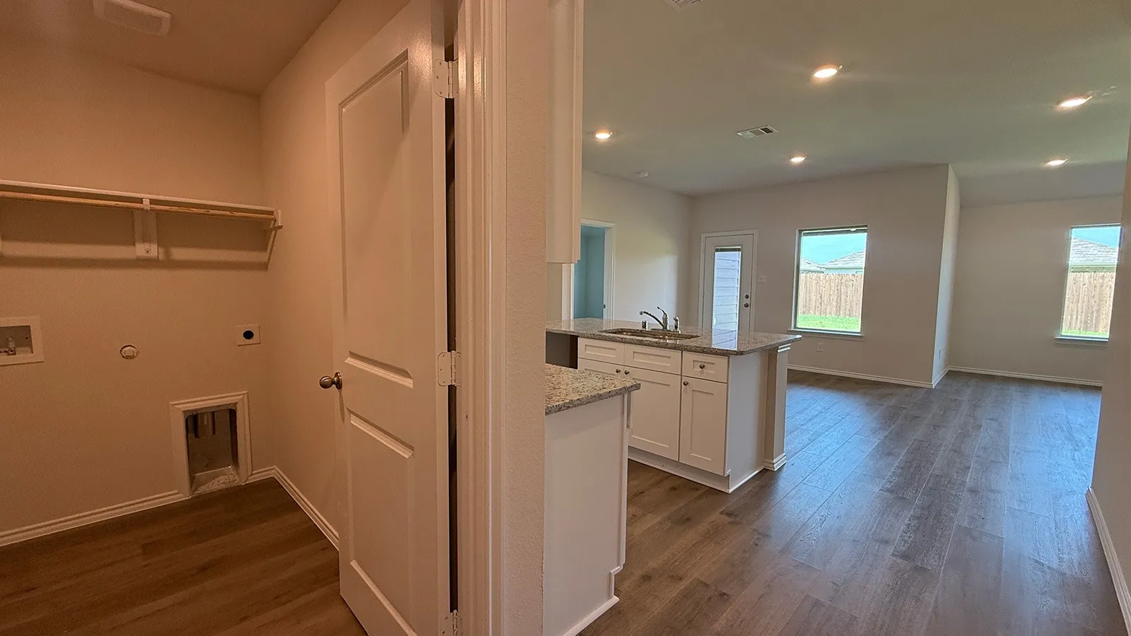Utility room with wood-finish flooring and built-in shelving, opening to an open-concept living space with recessed lighting, a kitchen island with sink, and light-colored countertops