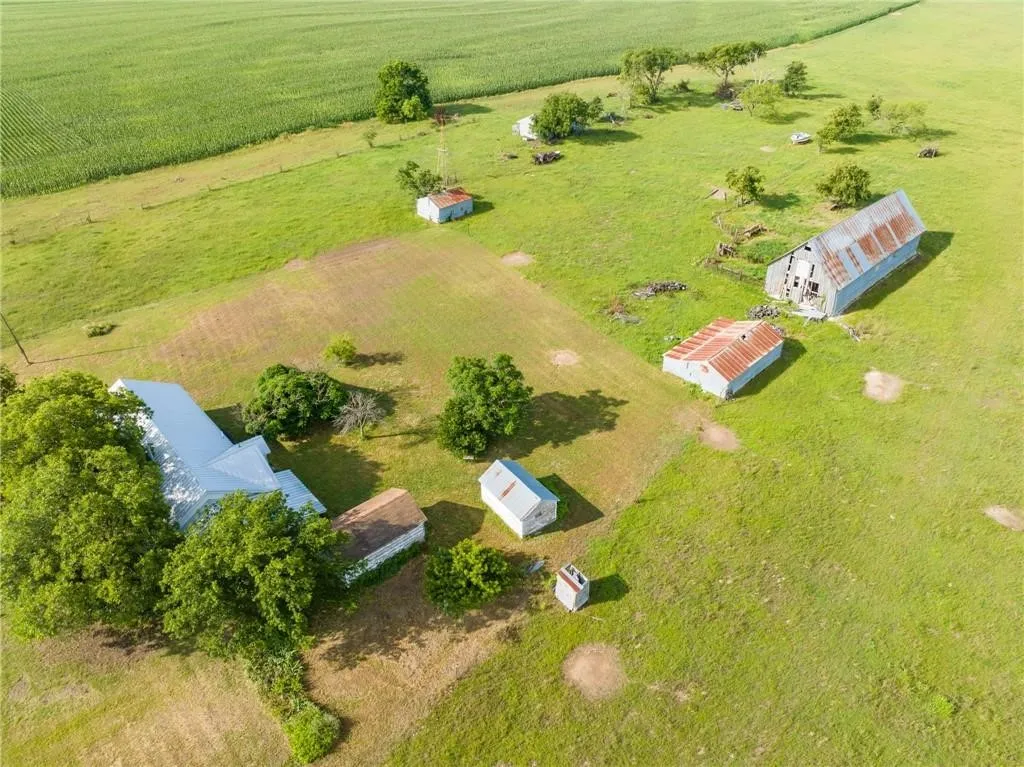 Expansive green acreage featuring multiple buildings, including a main building with a white roof, several smaller structures with weathered metal roofs, and a larger barn with a weathered metal roof