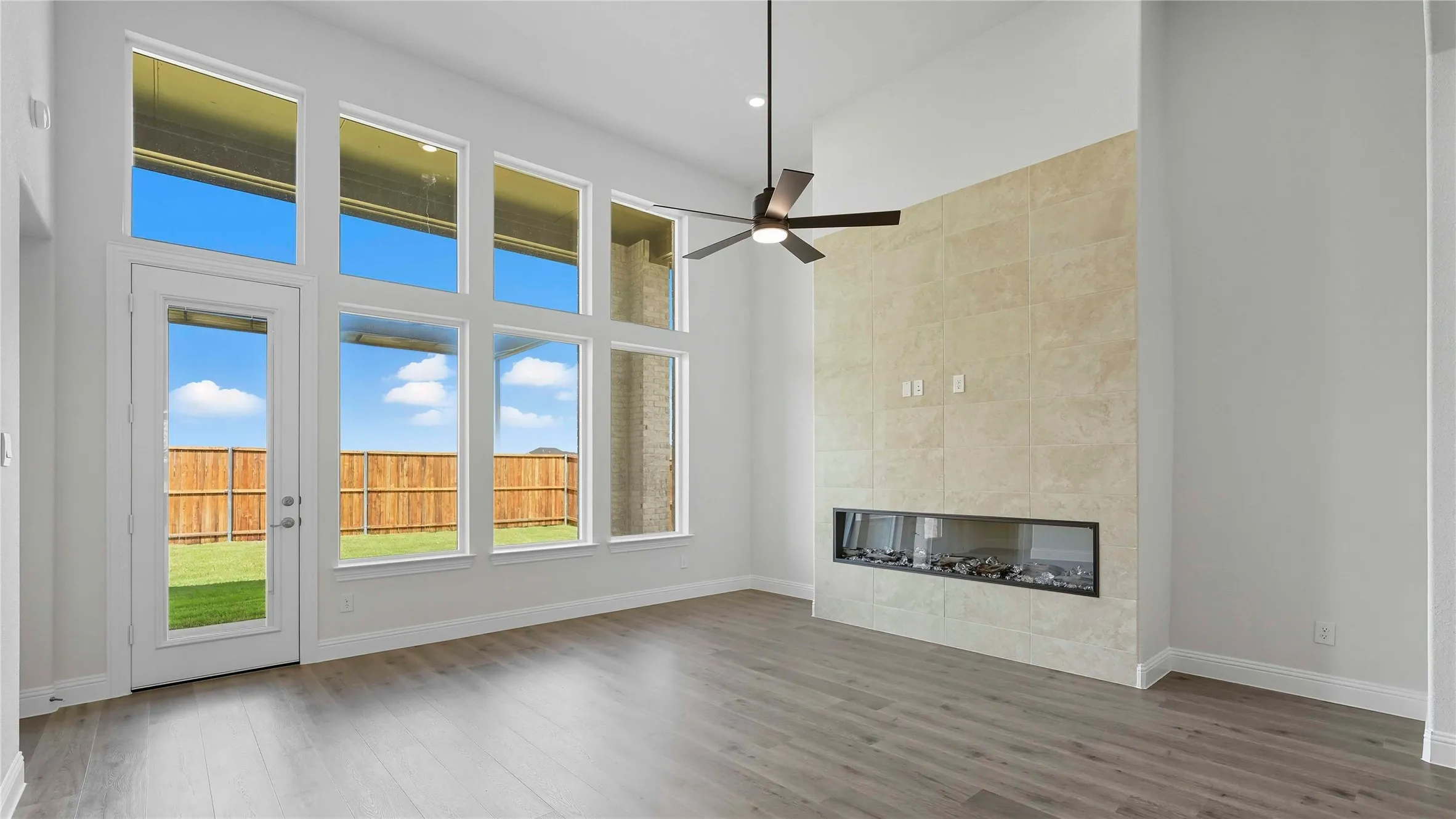 Spacious room featuring soaring ceilings, a full wall of tall windows and a glass-paneled exterior door, a sleek linear fireplace with a textured tile surround, wood-finish flooring, and a modern ceiling fan
