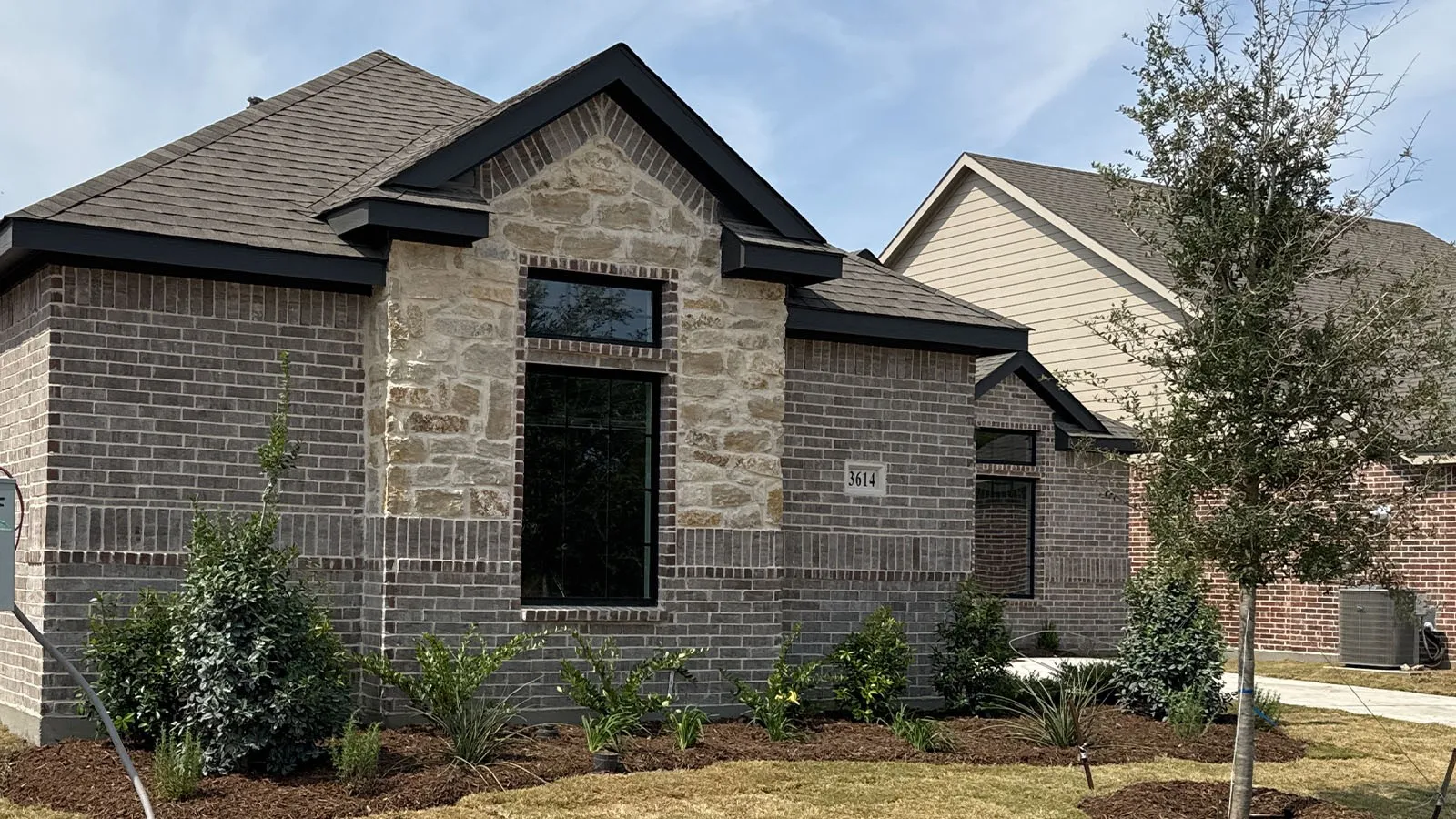 Exterior facade featuring a combination of brick and stone masonry, dark-framed windows, a dark shingle roof, and landscaped grounds with mature plantings