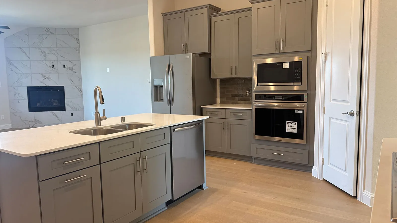 Open-concept kitchen with light wood-finish flooring, gray shaker cabinetry, stainless steel appliances, and a kitchen island featuring a double-basin sink
