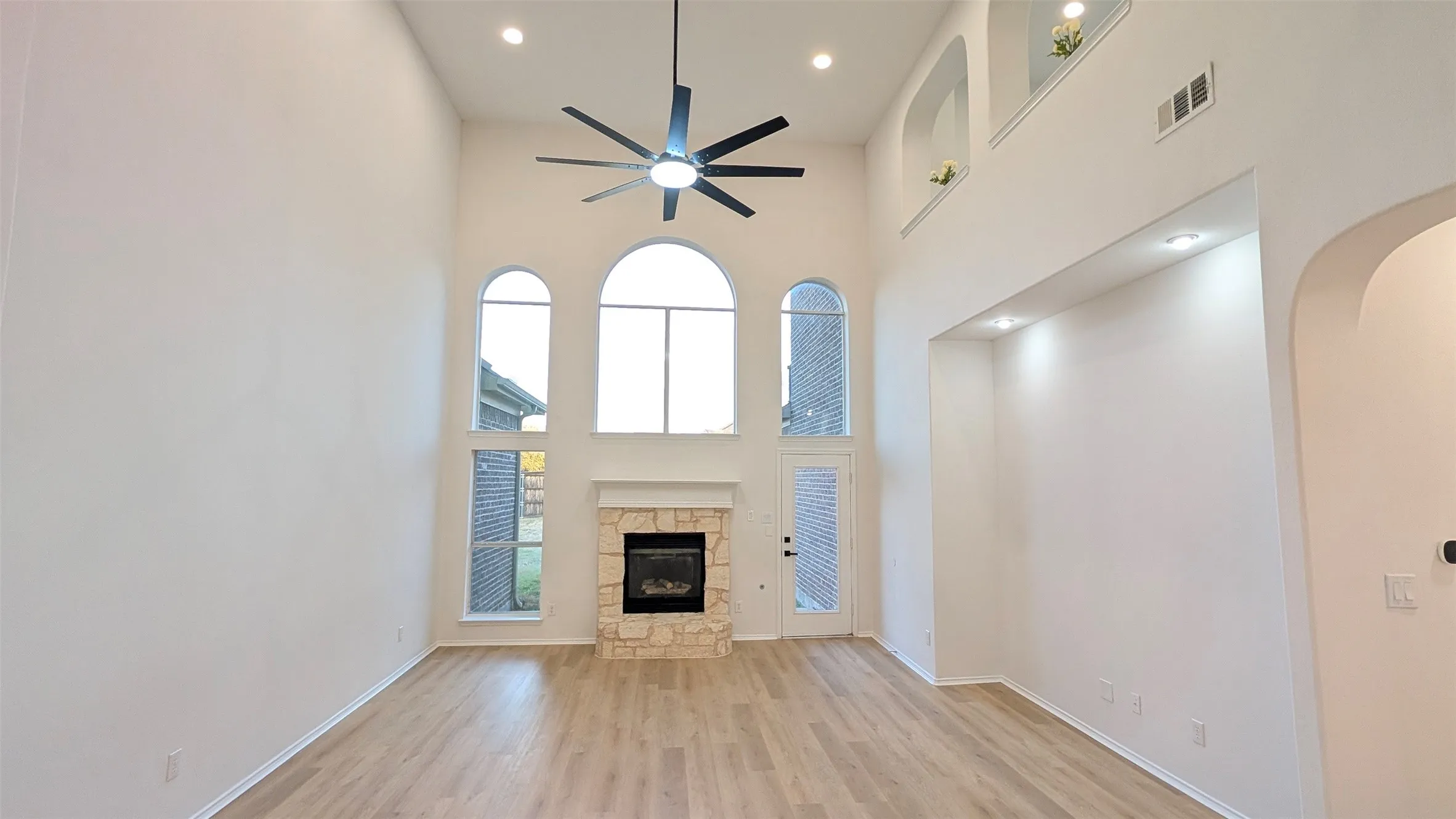 Two-story living area featuring a stone fireplace with a white mantel, arched windows, and light wood-finish flooring
