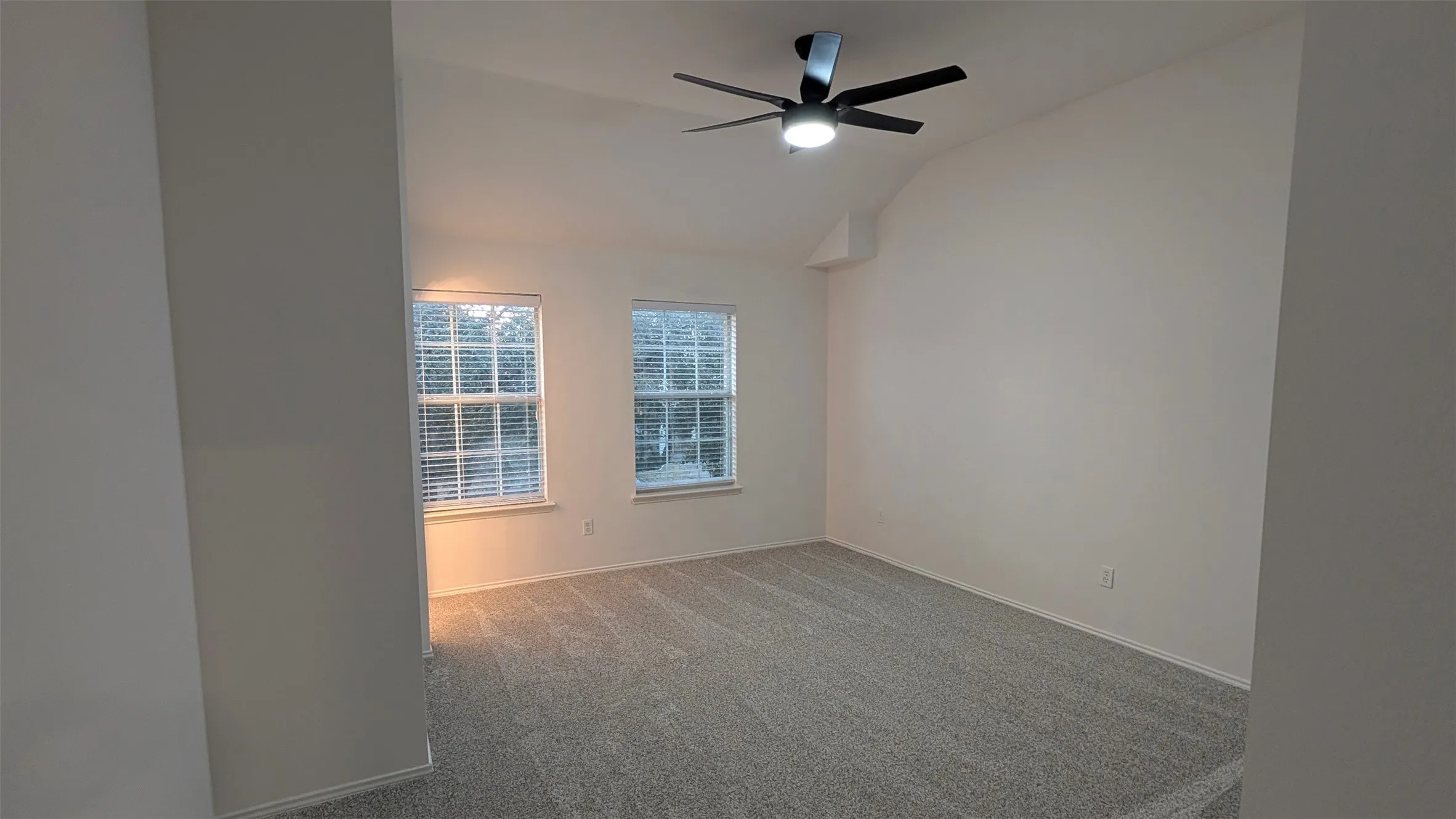 Spacious room featuring vaulted ceilings, light gray carpeting, two windows with blinds, and a modern ceiling fan with integrated lighting
