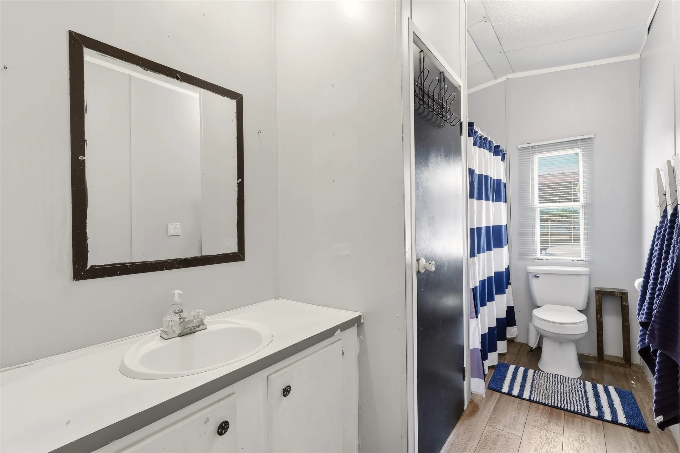 Bathroom featuring a white vanity with an integrated sink, a rectangular framed mirror, wood-finish flooring, a toilet, and a window with blinds