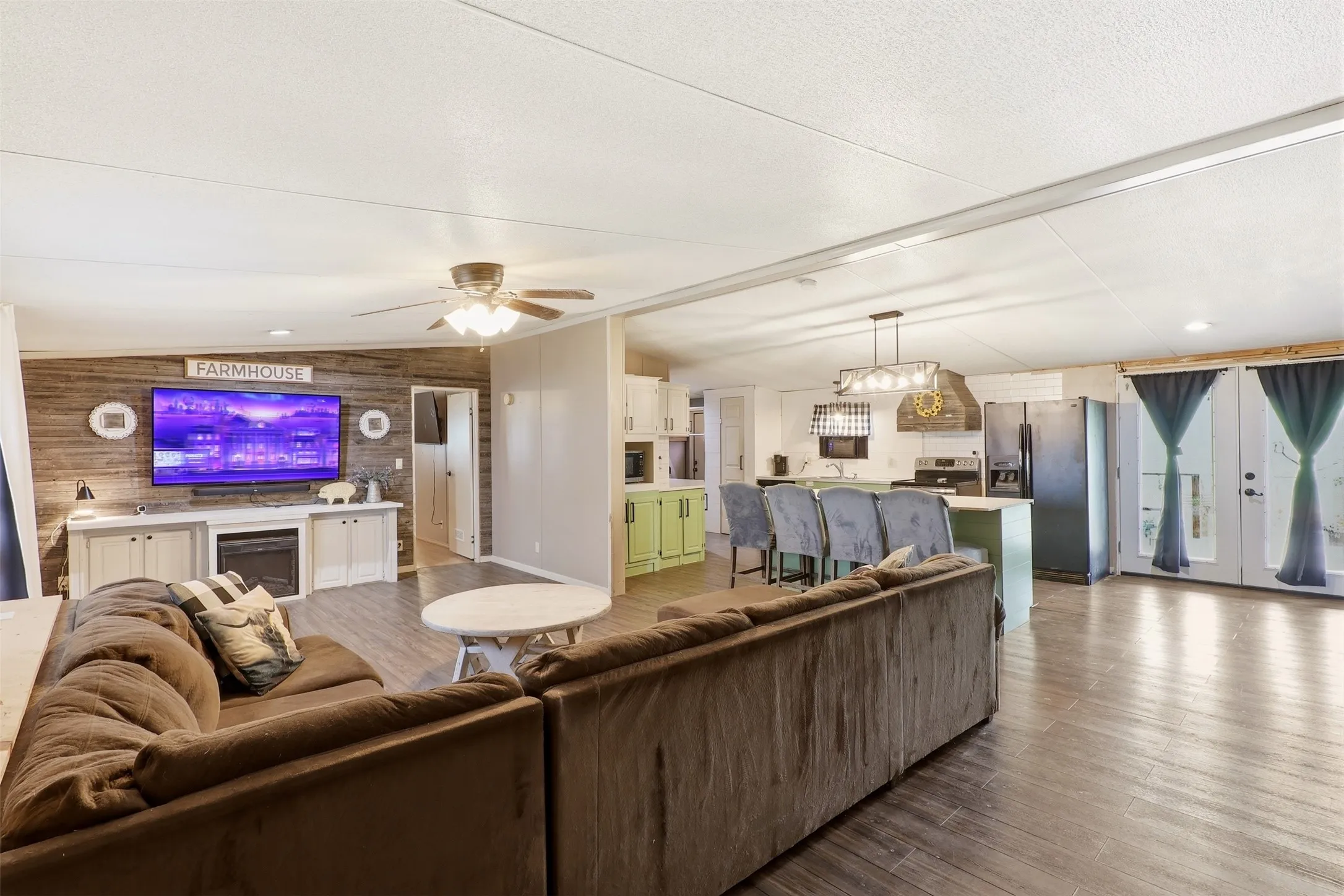 Open-concept living area featuring wood-finish flooring, a shiplap accent wall, built-in media console with an electric fireplace, a ceiling fan, and French doors