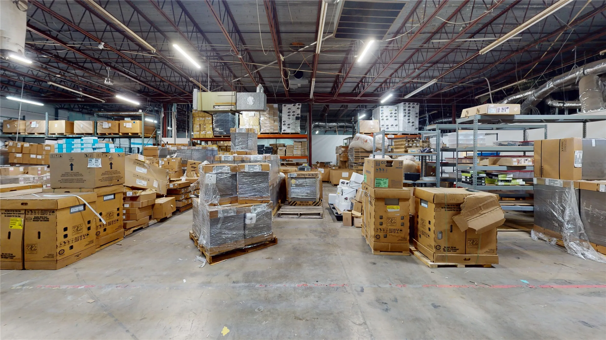 Spacious industrial warehouse featuring a concrete floor, exposed metal beam ceiling structure, fluorescent lighting, and extensive shelving units