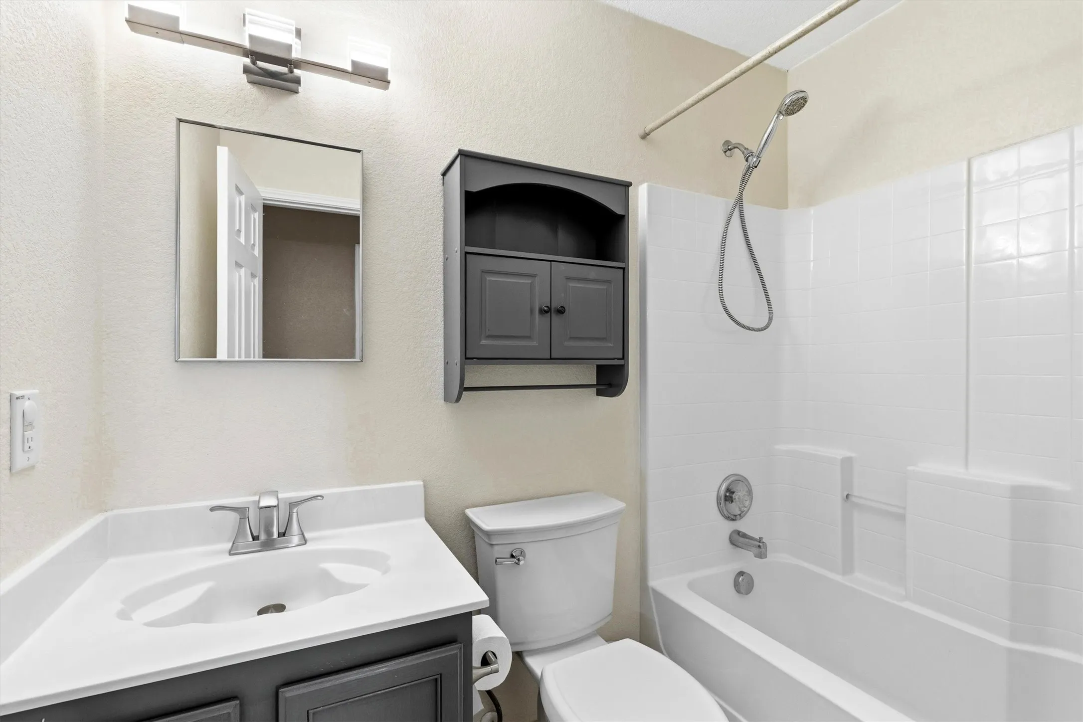 Bathroom featuring a white bathtub with shower, white tile-patterned surround, a dark gray vanity with an integrated white sink and countertop, and a wall-mounted storage cabinet