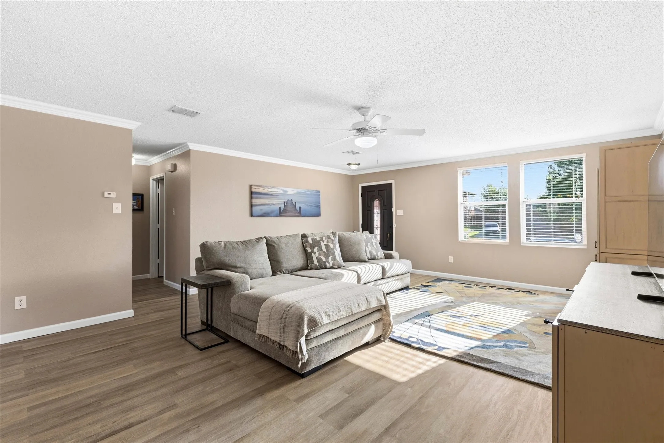 Spacious living area featuring wood-finish flooring, two large windows, a ceiling fan with integrated lighting, a dark paneled entry door, and crown molding