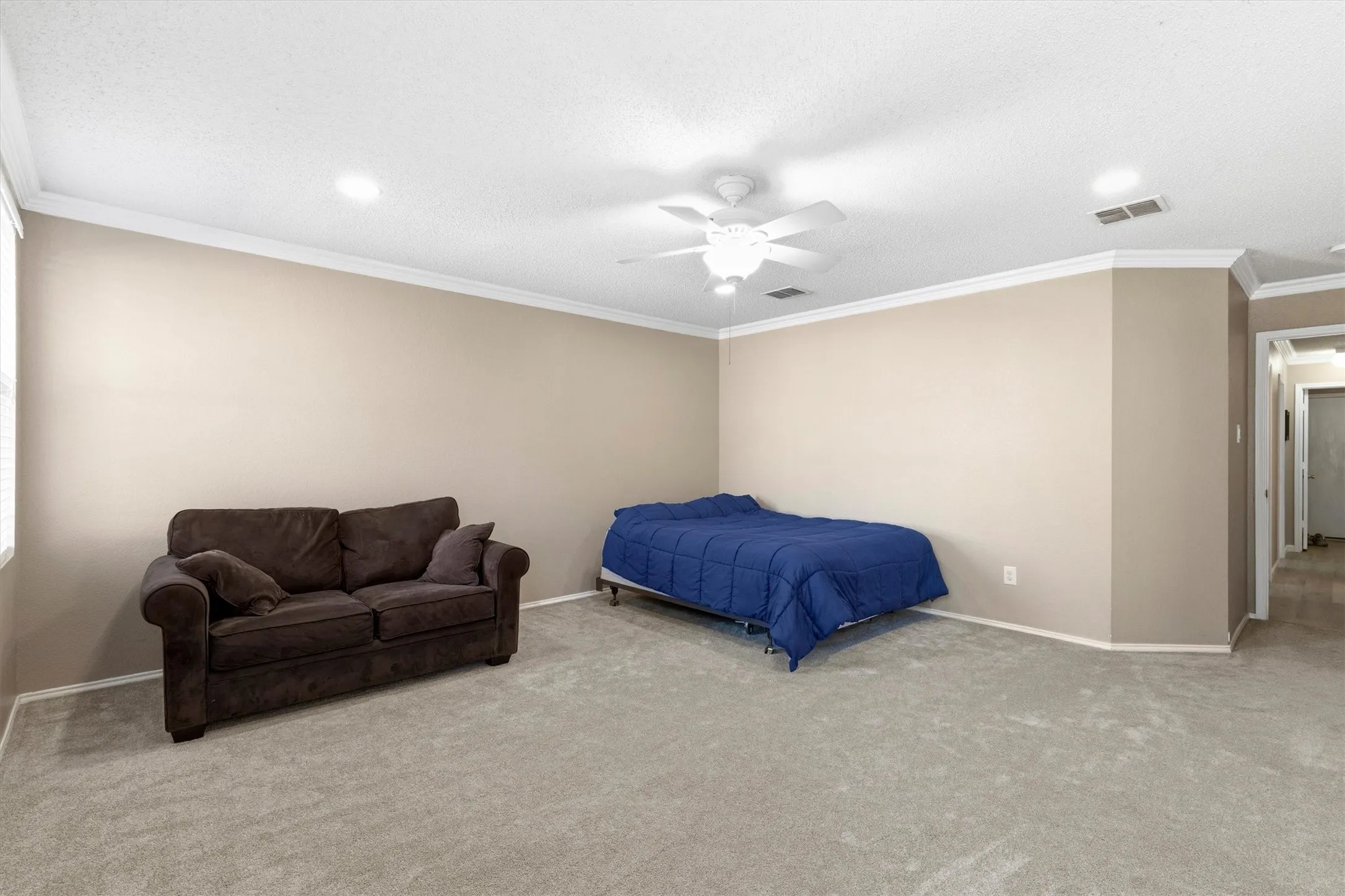 Spacious room featuring neutral carpeting, a ceiling fan with light, recessed lighting, crown molding, and a hallway entry