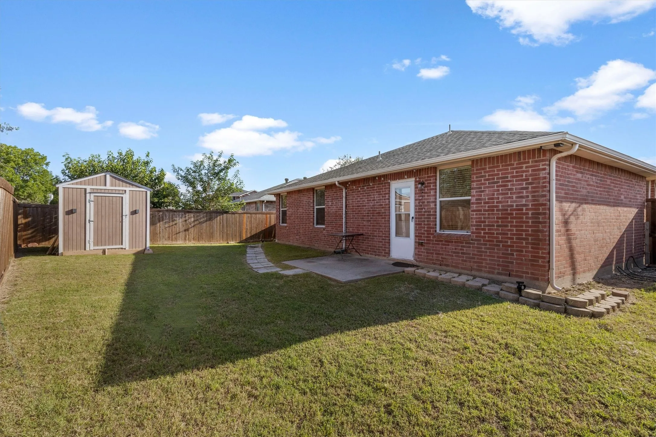 Spacious backyard featuring a brick exterior, a concrete patio, a detached storage shed, a wood fence, and a stone garden path