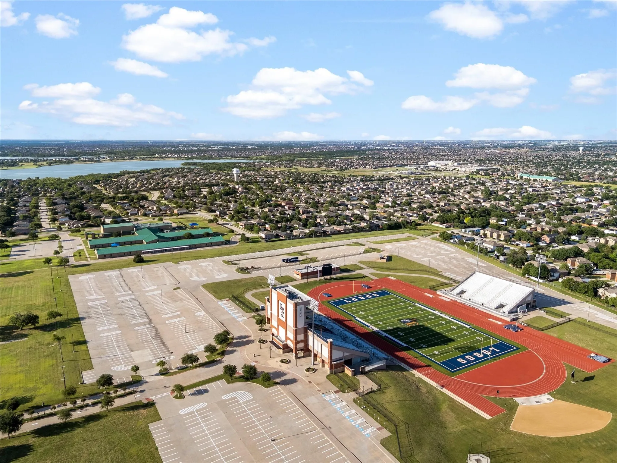 Aerial perspective showcasing a large athletic complex with a football field and track, surrounded by extensive asphalt parking areas