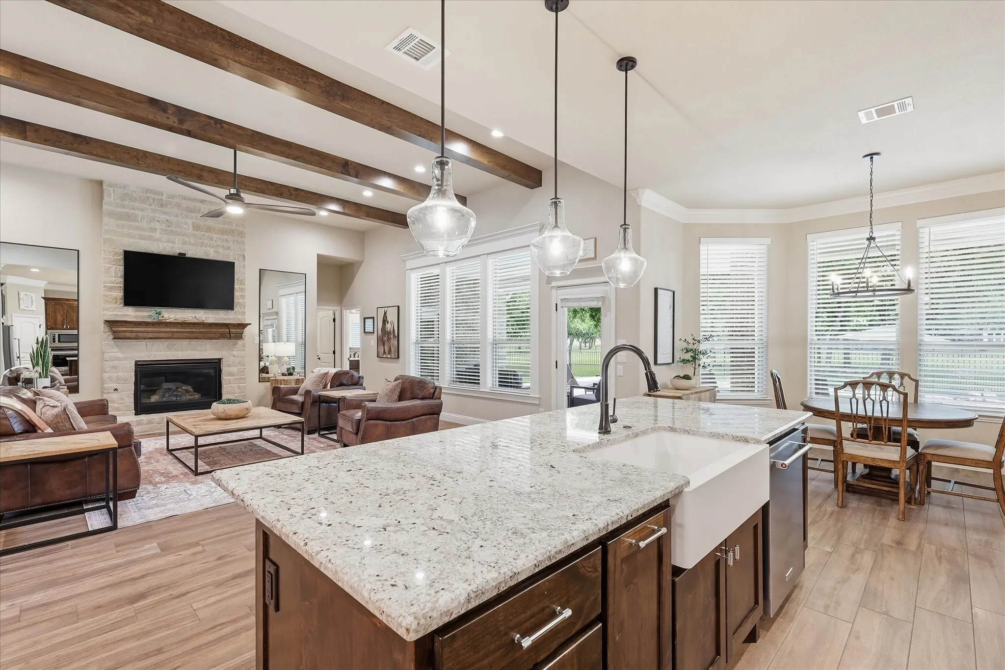 Spacious open-concept living area featuring a kitchen island with stone countertops and a farmhouse sink, wood-finish flooring, exposed ceiling beams, a stone fireplace, and multiple windows
