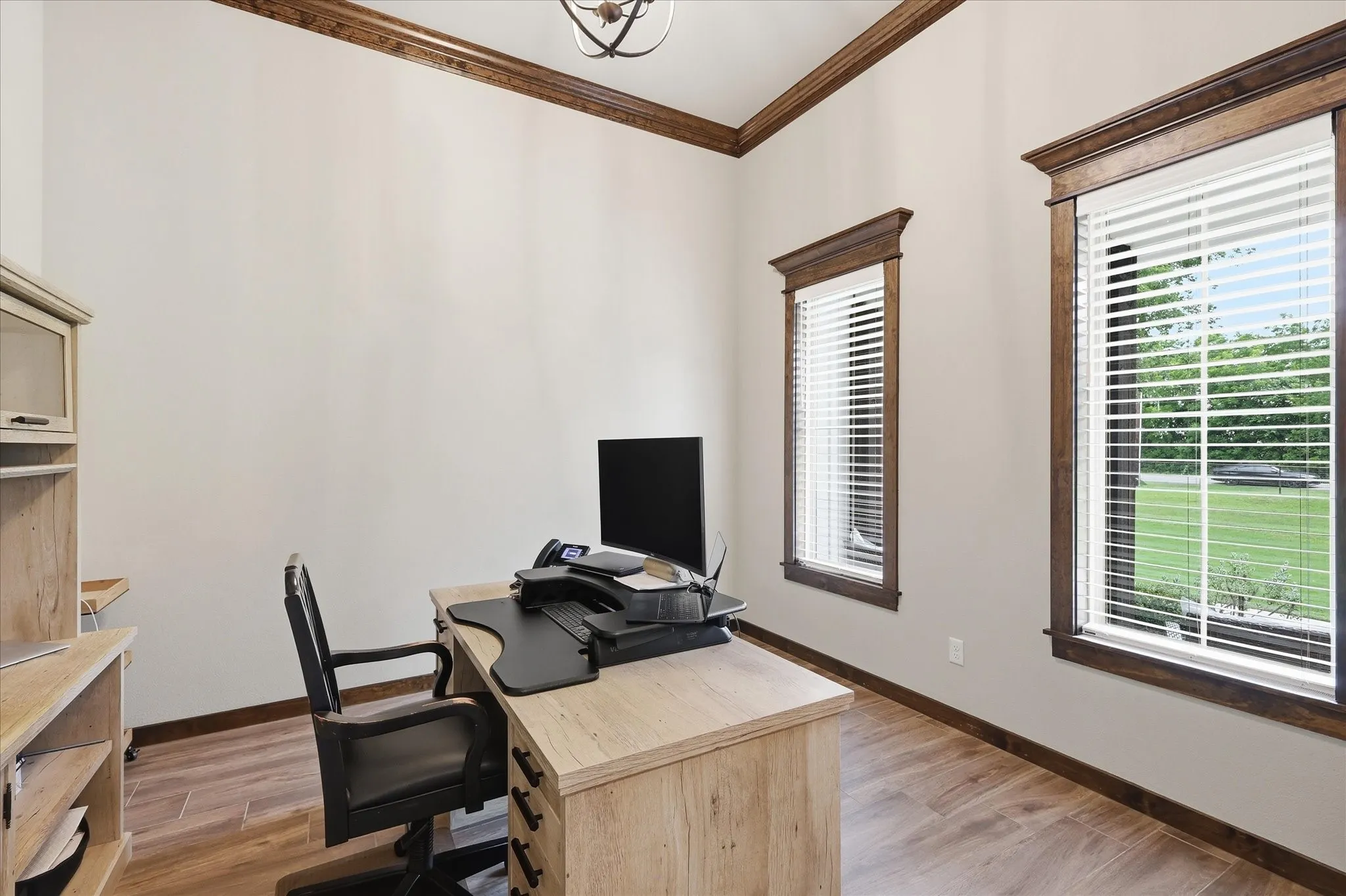 Room with wood-finish flooring, two large windows with blinds, detailed dark wood trim, crown molding, and a visible portion of a decorative ceiling light fixture
