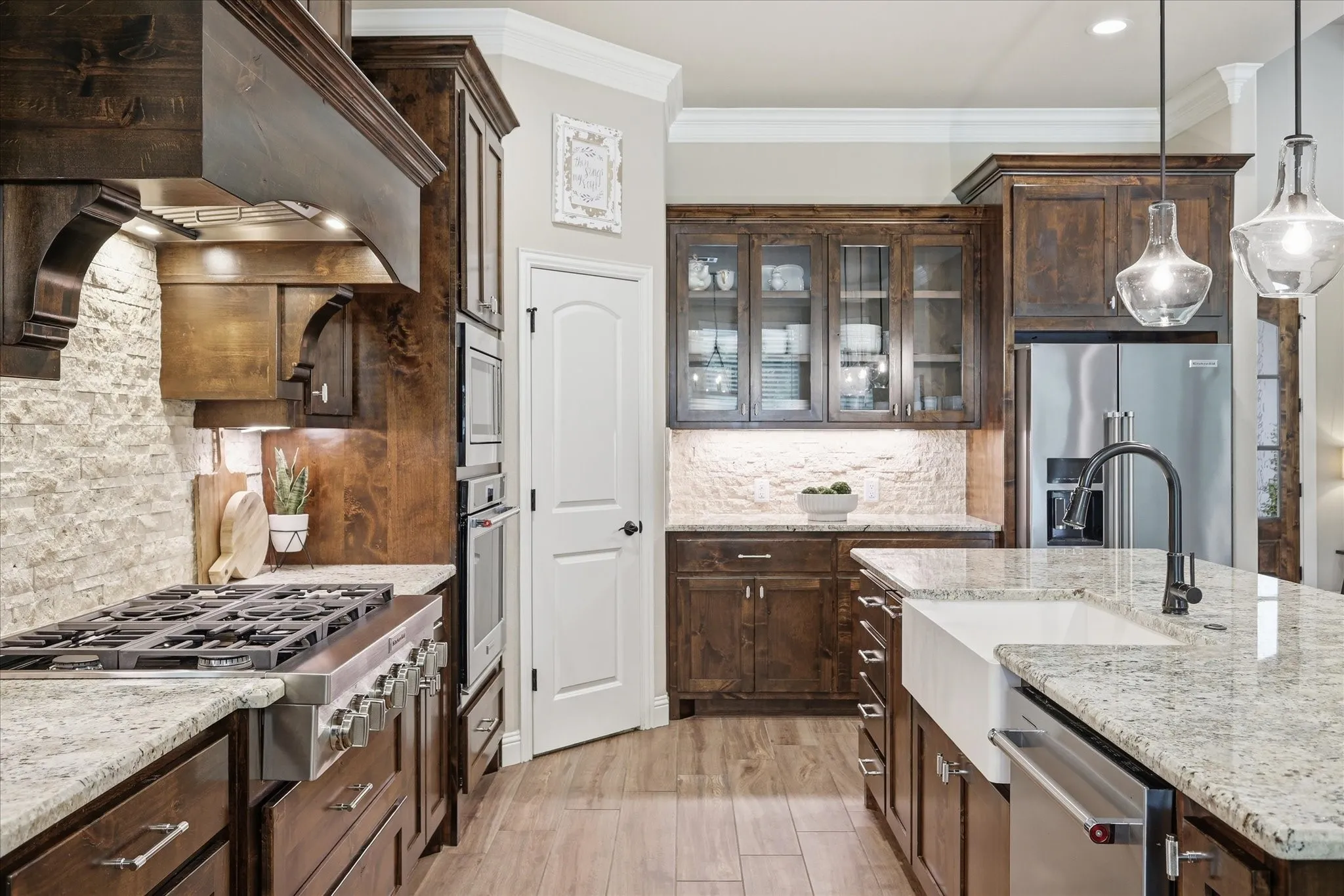 Gourmet kitchen featuring rich wood cabinetry, stone countertops, a large center island with a farmhouse sink, stainless steel appliances, and a stacked stone backsplash
