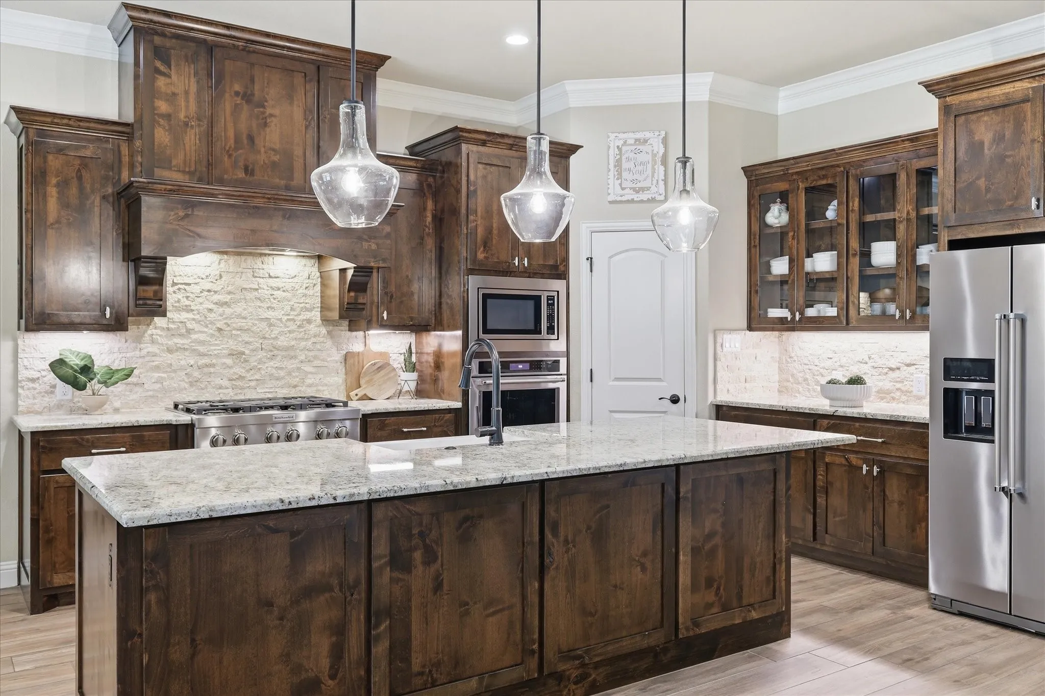 Gourmet kitchen featuring a large central island with light granite-finish countertop, dark wood cabinetry, stainless steel appliances, and a stacked stone backsplash
