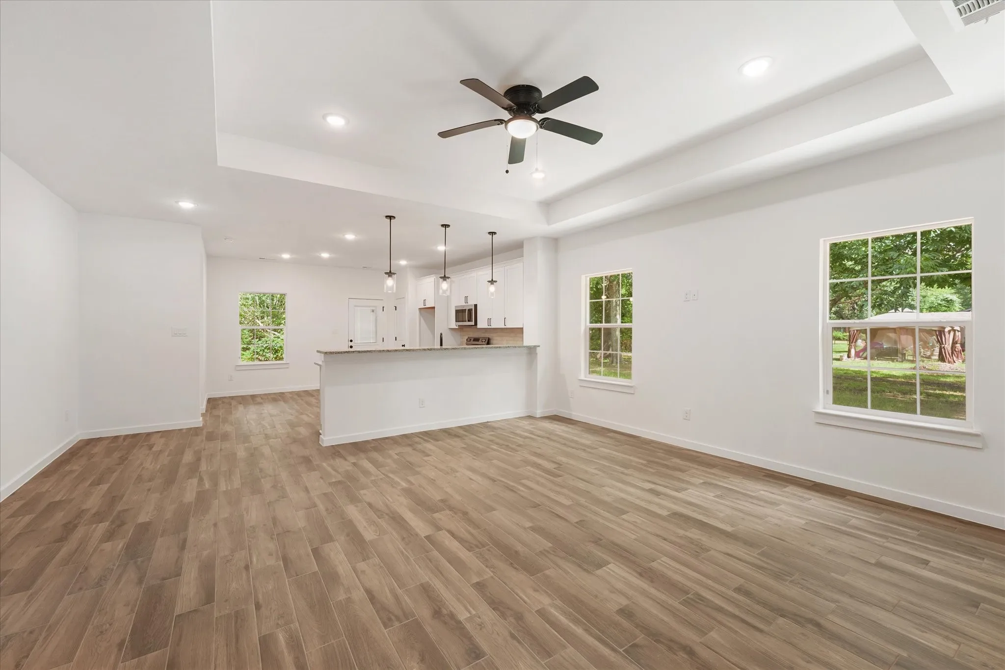 Spacious living area featuring wood-finish plank flooring, white walls, and a tray ceiling with a ceiling fan
