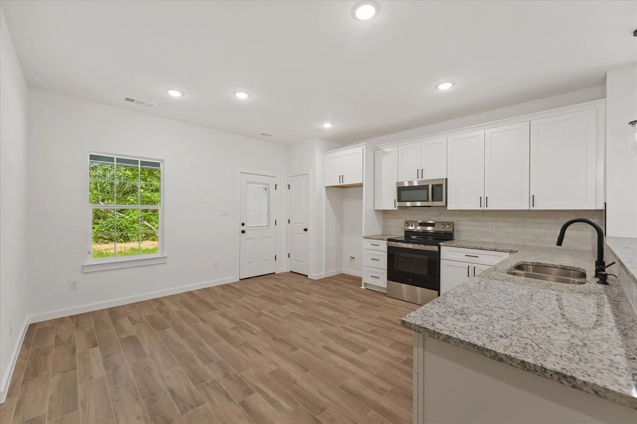 Open-concept kitchen and living area featuring wood-finish flooring, white shaker cabinetry, stainless steel appliances, granite countertops, and recessed lighting