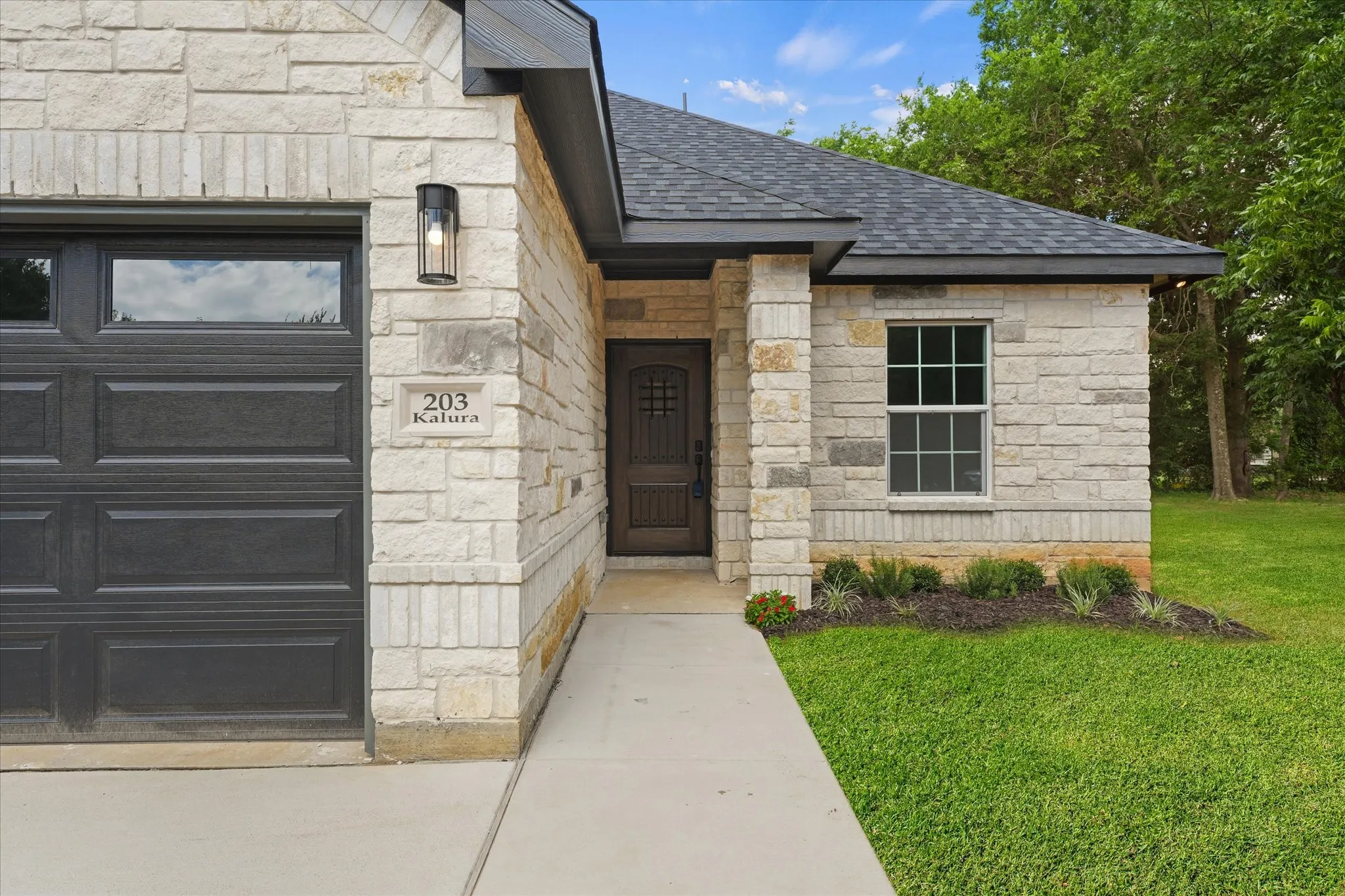 Stone veneer exterior featuring a dark paneled garage door, a front entry door with decorative glass, an exterior sconce light, and a manicured lawn with planted landscaping