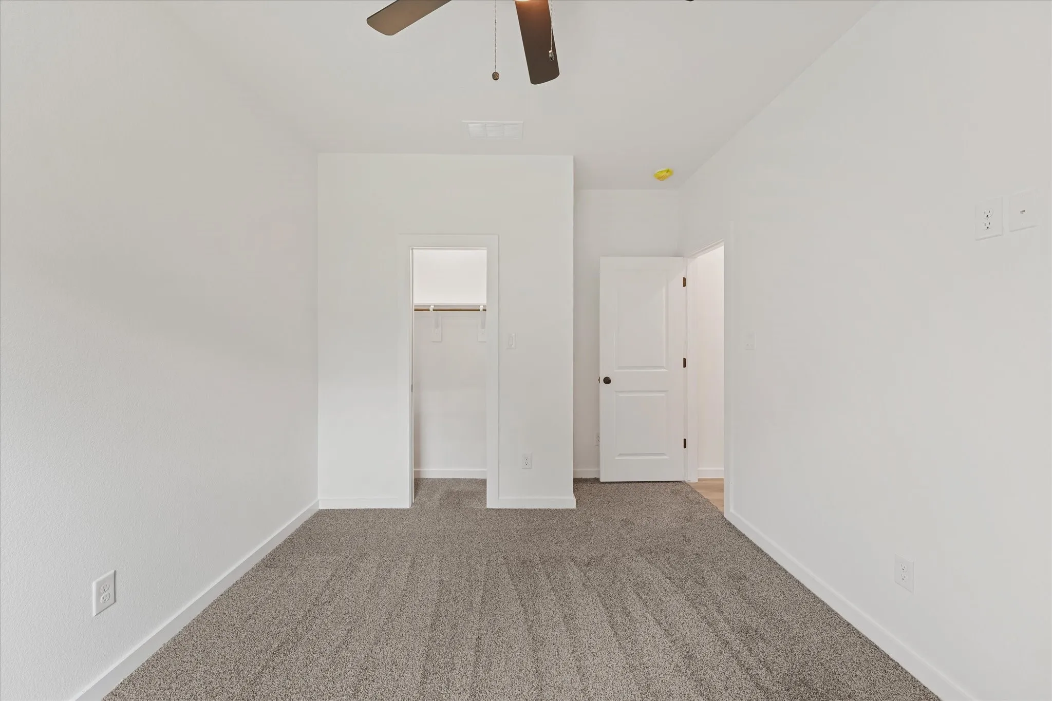 Interior room featuring plush gray carpeting, white painted walls, a built-in closet with shelving, a ceiling fan, and a white interior door with dark hardware