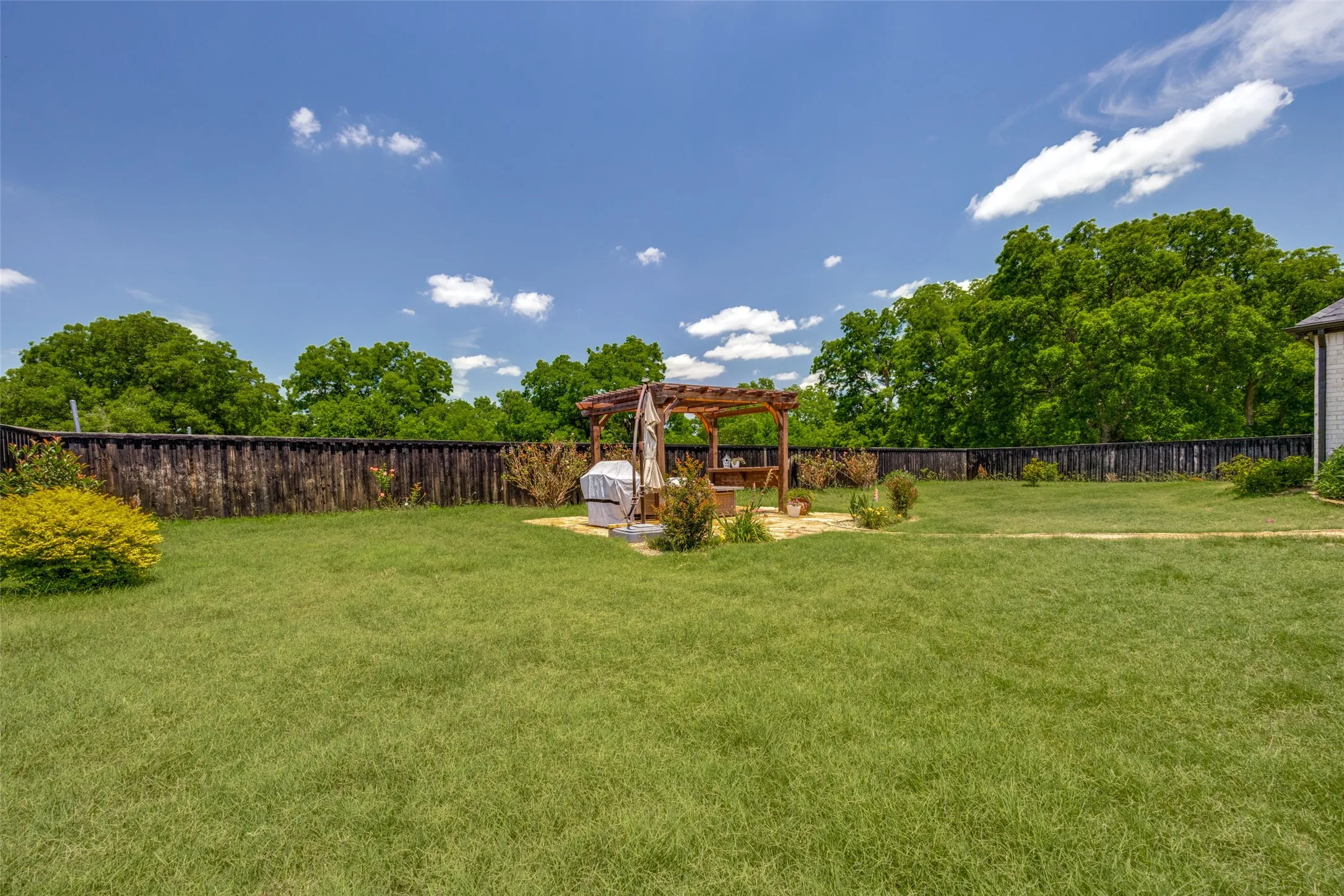 Expansive grassy backyard featuring a wooden pergola on a light-colored patio, mature privacy fencing, and established perimeter trees