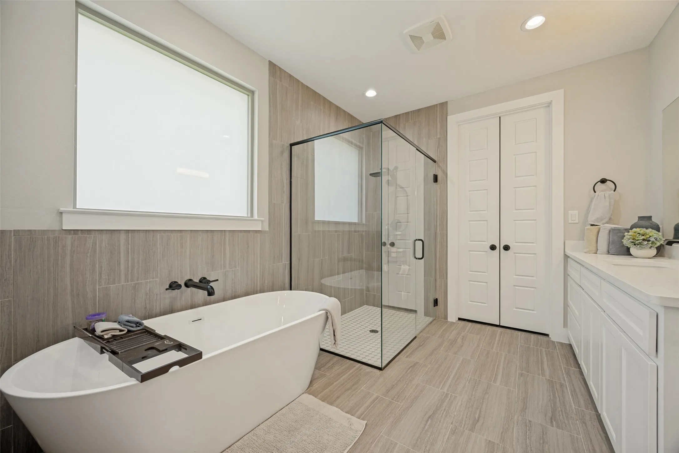 Contemporary bathroom featuring a freestanding soaking tub with wall-mounted matte black fixtures, a spacious glass-enclosed shower with tiled walls and floor, and a white double vanity with a light-colored granite countertop